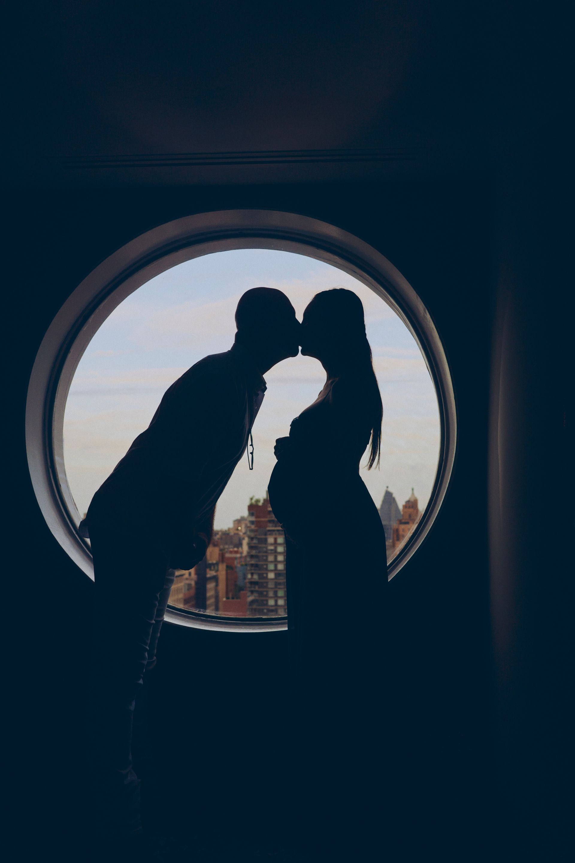 Silhouette of couple kissing, woman pregnant. Round window frames city skyline.