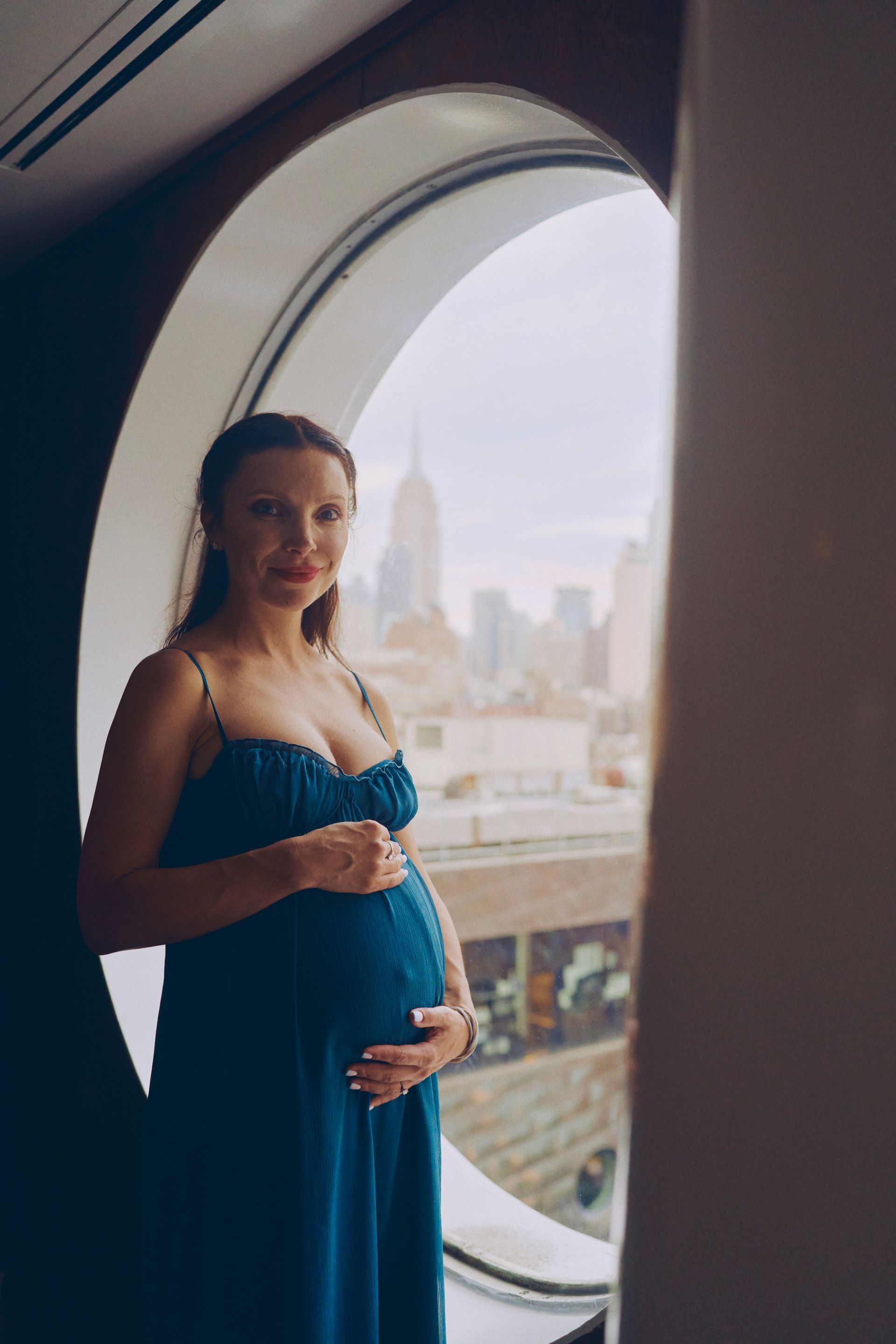 Pregnant person stands in water, sunlight shining. Wearing a white dress, hands on belly.