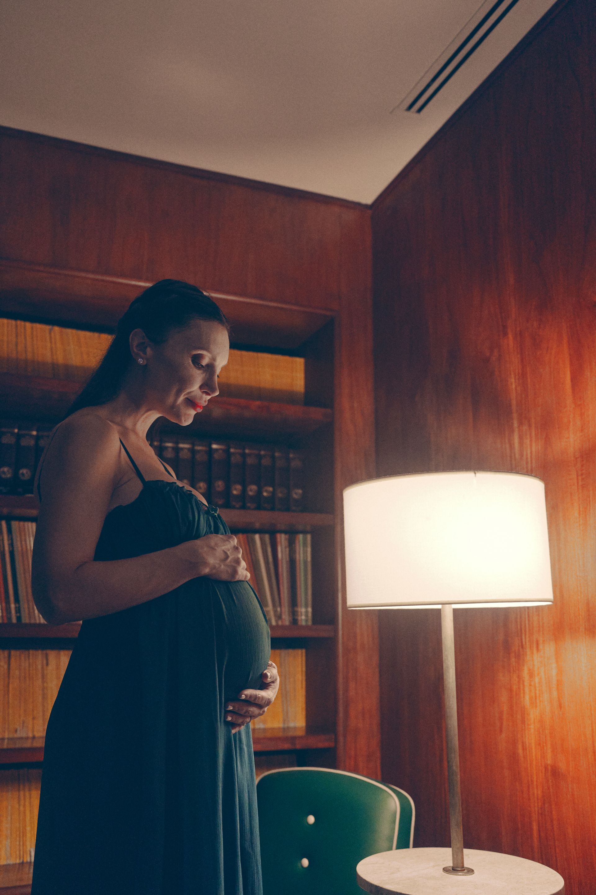 Pregnant woman in a dark blue dress, caressing her belly in a wood-paneled room with a lamp and bookcase.