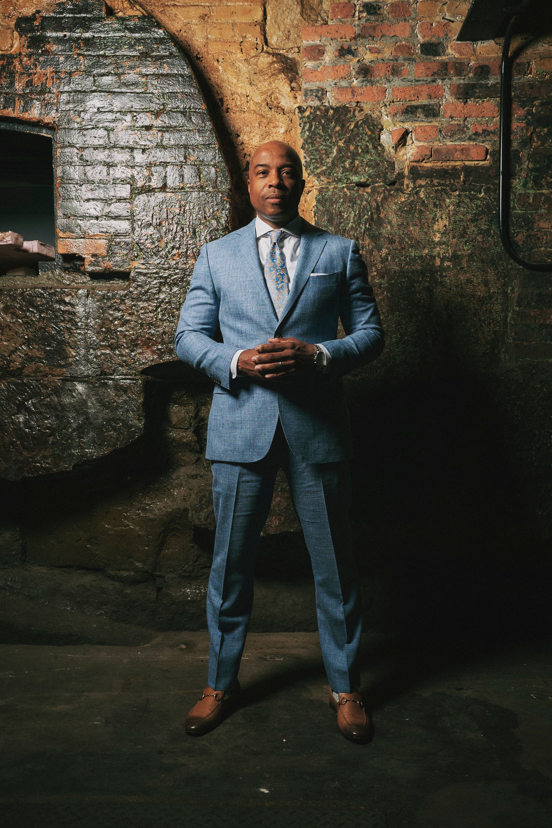 Man in blue suit stands in a brick-walled room, hands clasped, looking forward.