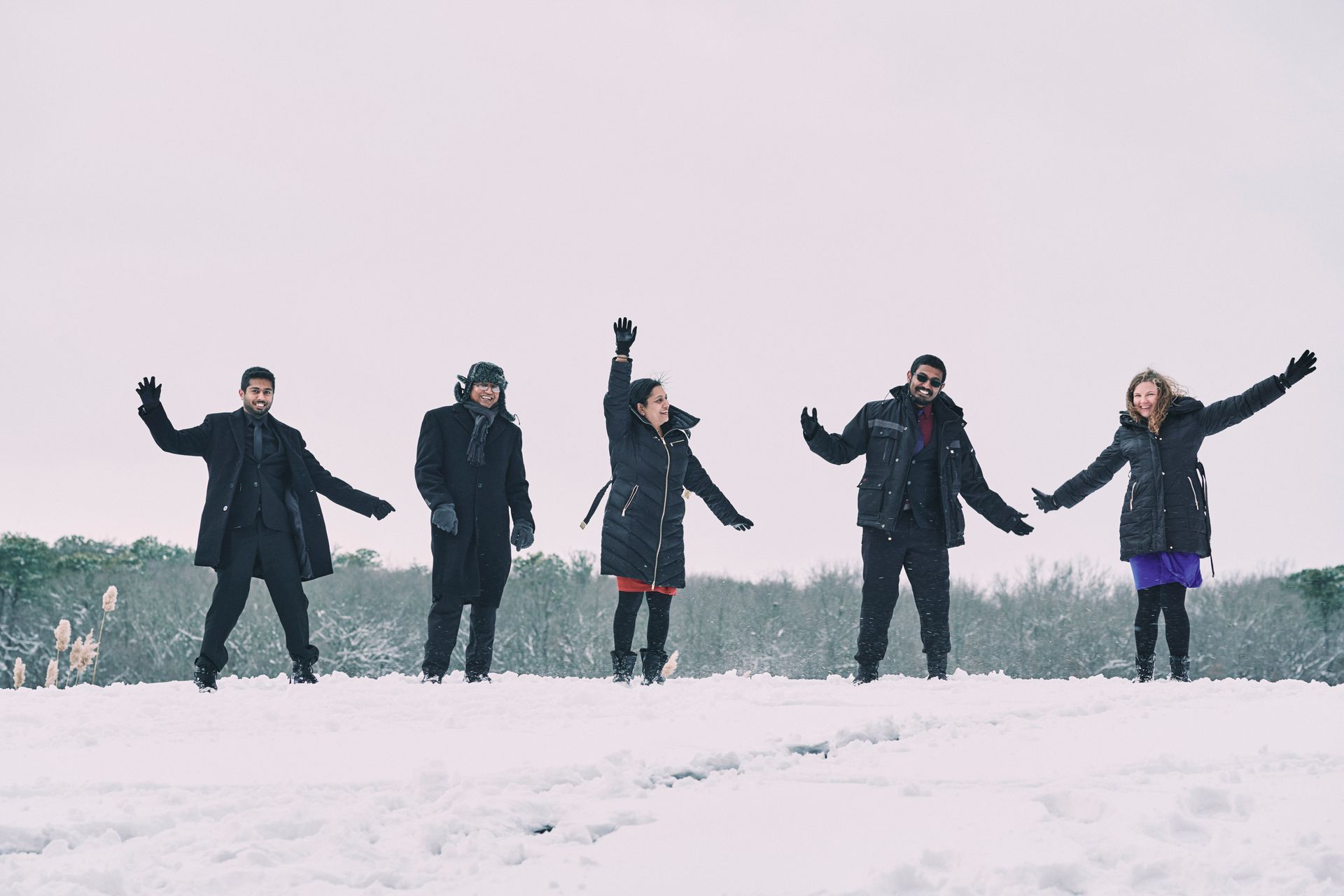Five people joyfully raise arms in a snowy field. They wear winter coats in a cloudy, overcast setting.