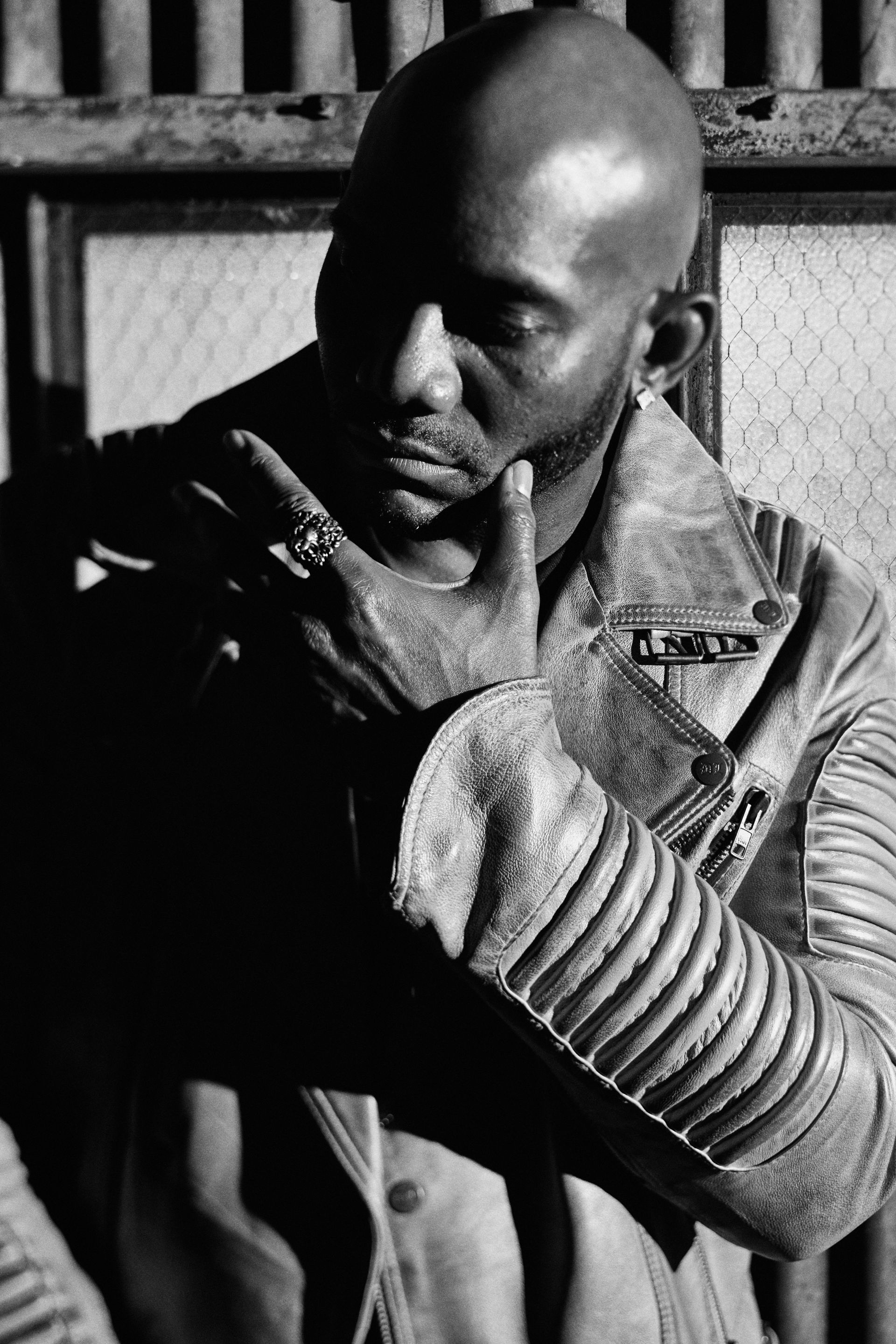 Bald Black man in leather jacket, hand on chin, thoughtful expression. Grayscale, industrial setting.
