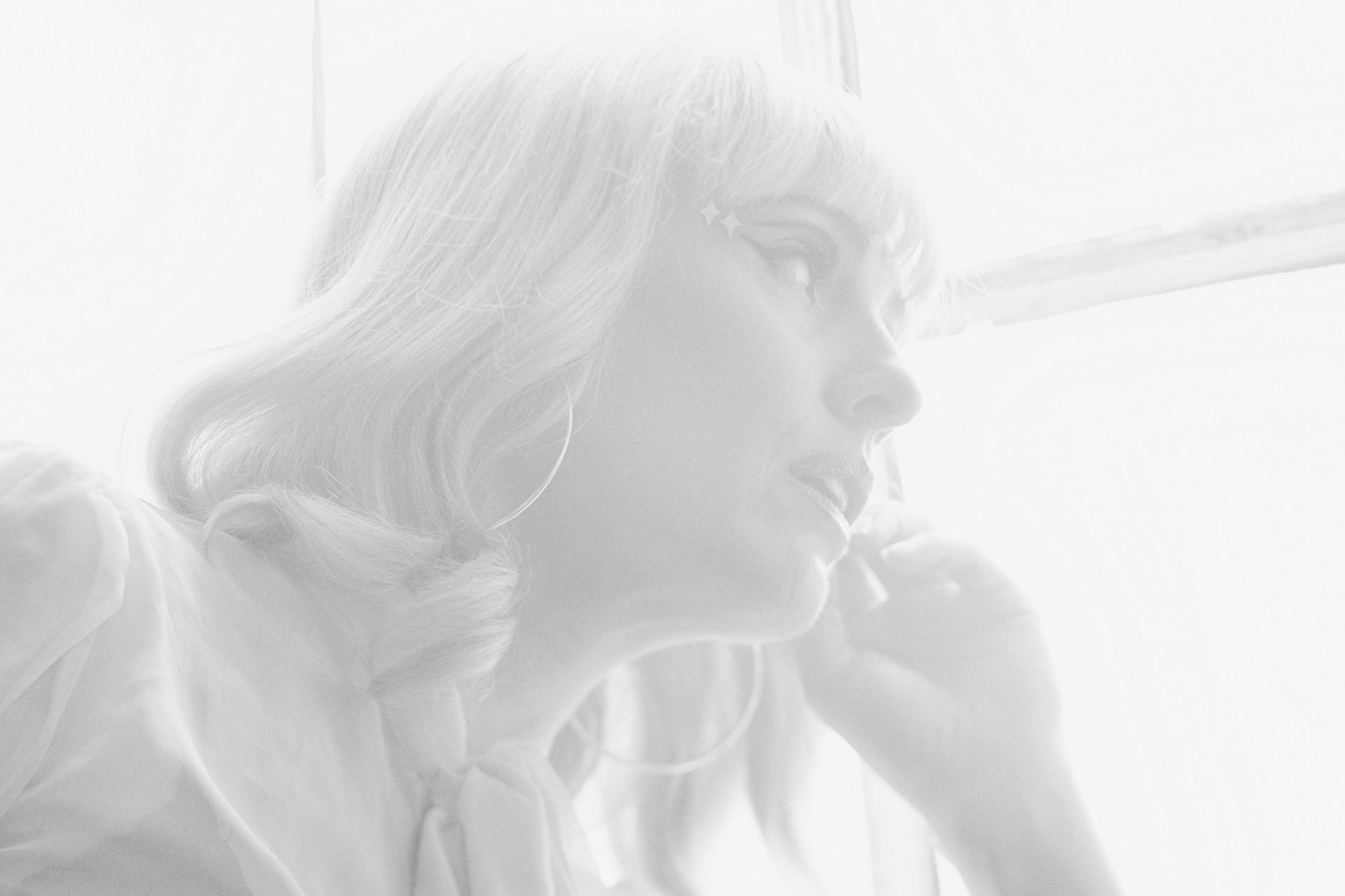 Blonde woman looking away, profile view, near a window, light background, wearing hoops.