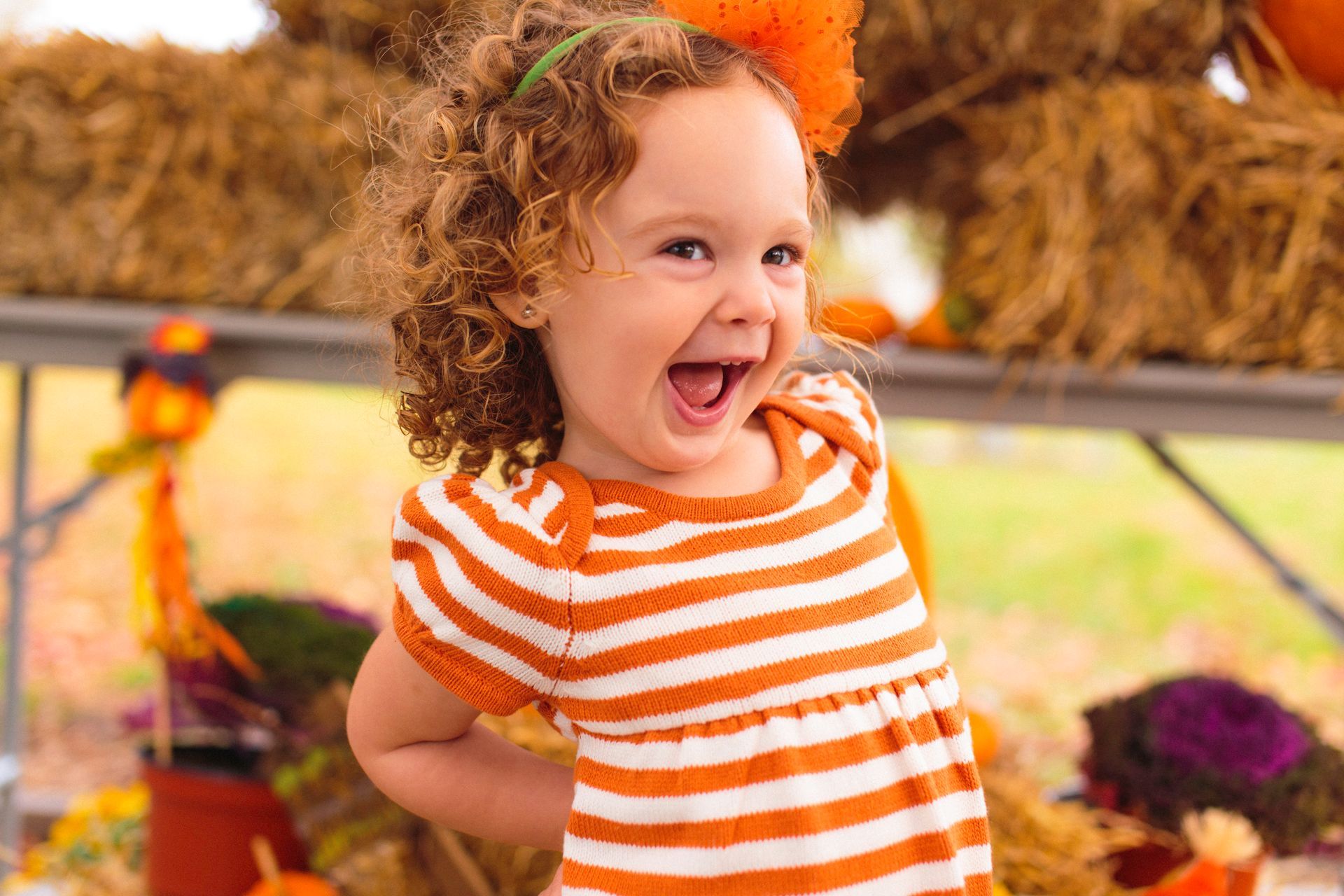 Young girl with curly hair laughs, wearing orange striped dress, with pumpkins and hay in background.