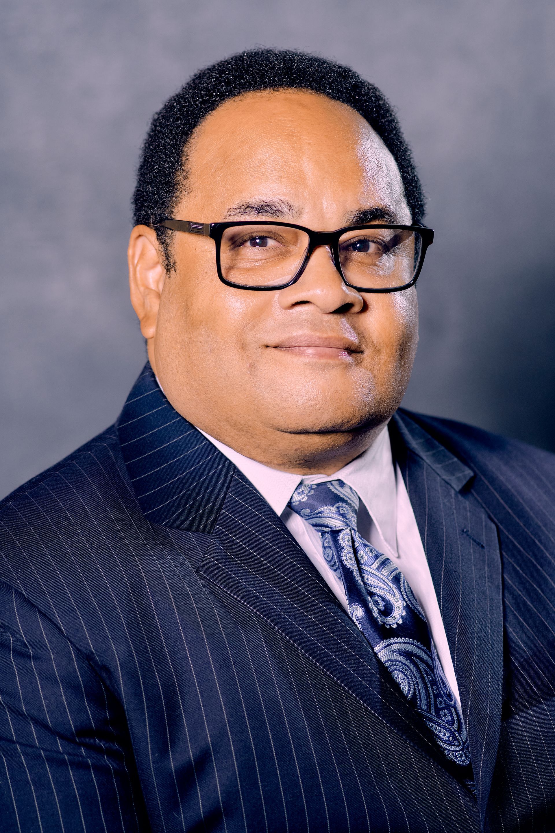 Man in a blue pinstripe suit, glasses, and paisley tie, smiling at the camera.