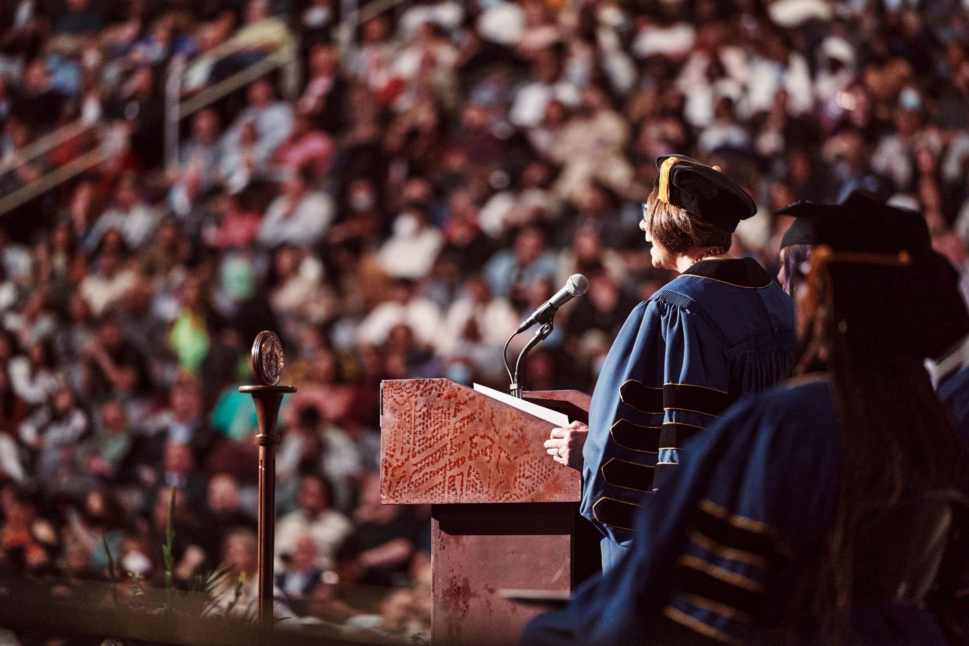 Graduation ceremony: Person in blue robes speaks at a podium, facing a large crowd.