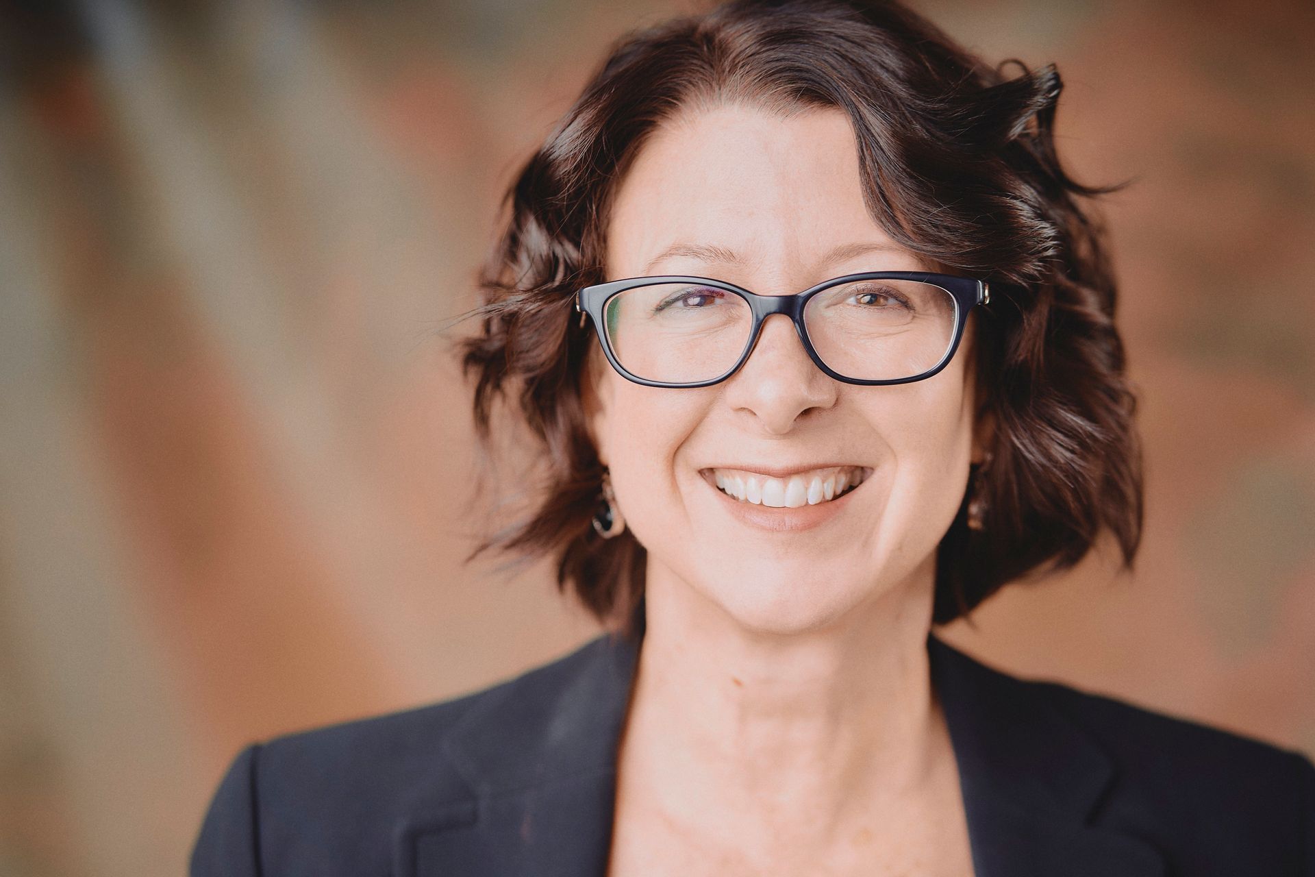 Woman with glasses, smiling, wearing a black blazer. Background: blurred colors.