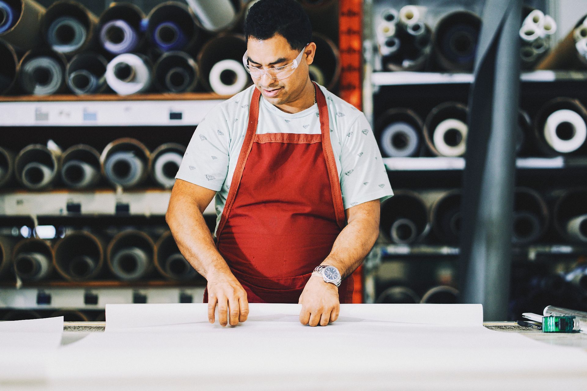 Man in red apron measures fabric in a workshop with rolls of material in the background.