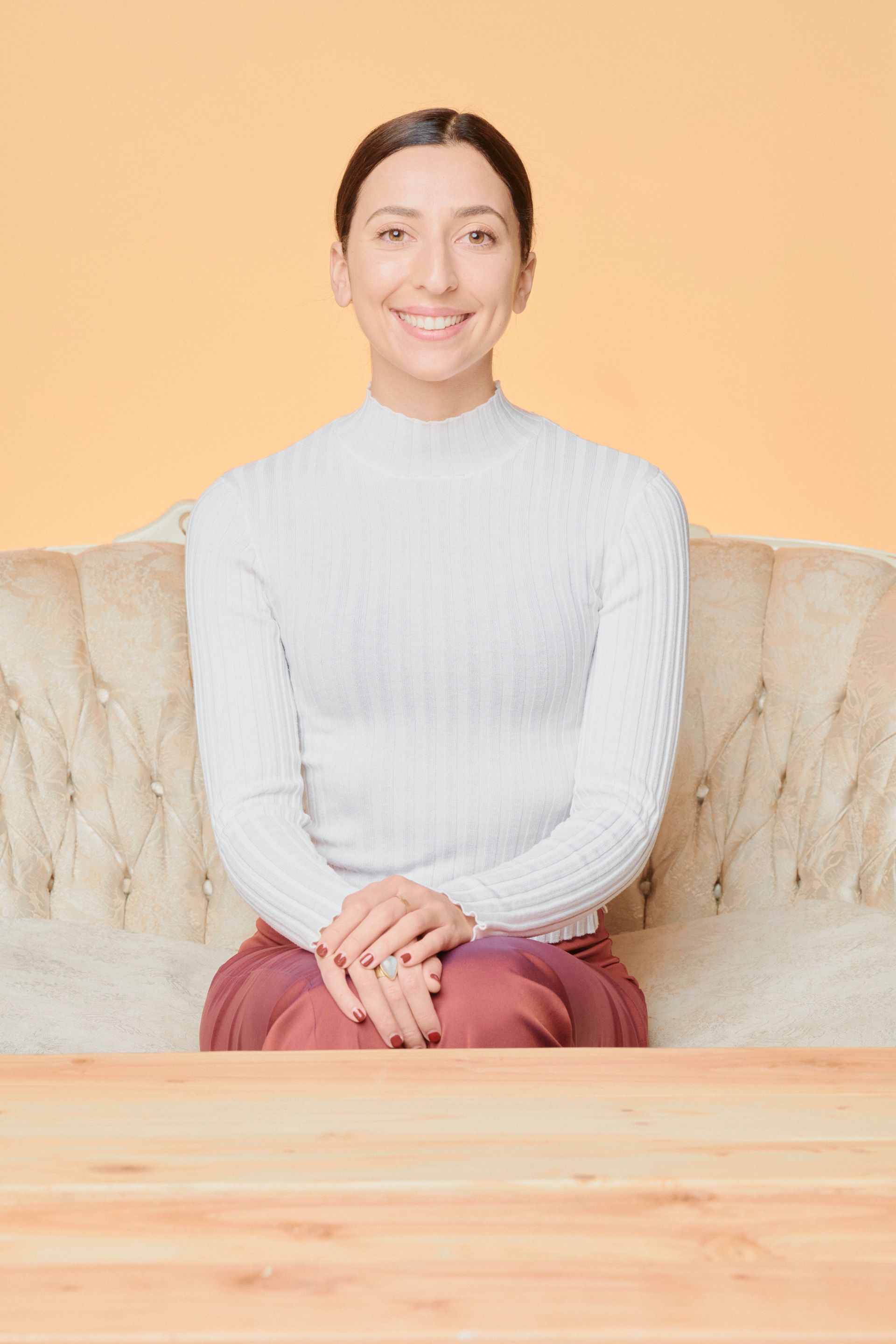 Woman seated on a cream-colored couch, smiling. Wearing white sweater and red pants. Neutral background.