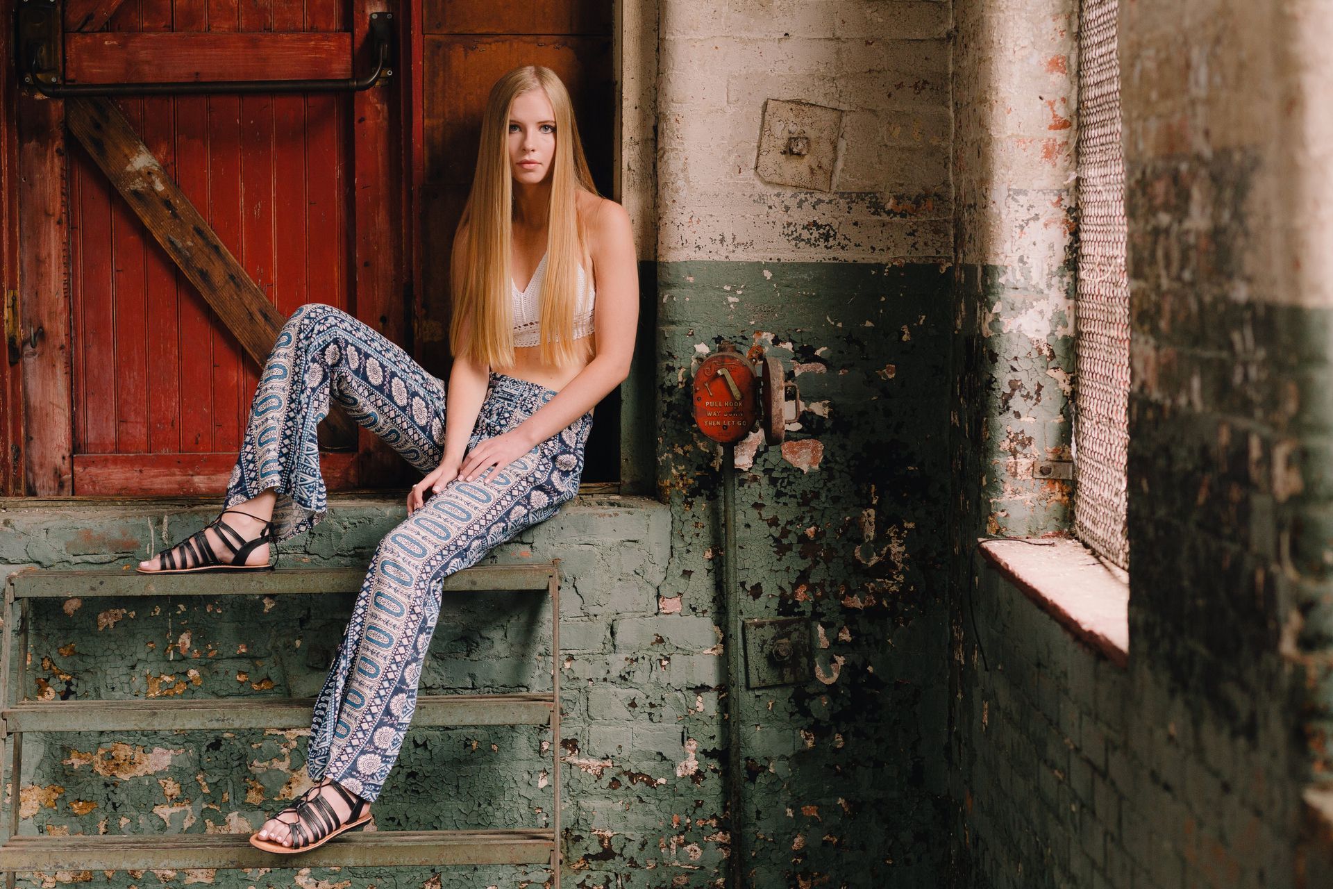 Woman with long blonde hair poses on steps, wearing printed pants, a white top, and sandals in a worn, rustic setting.