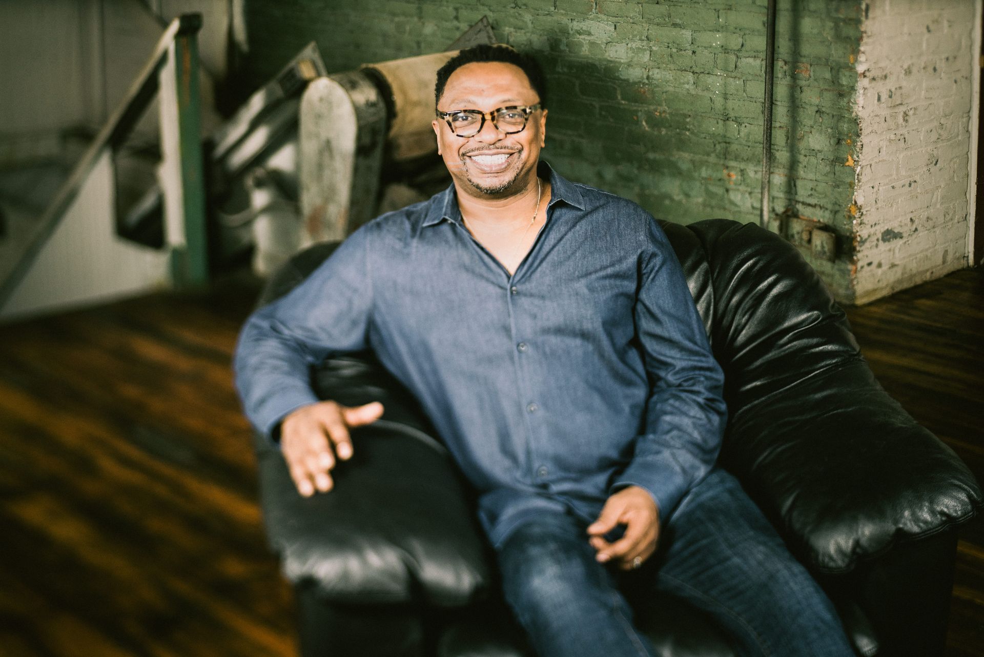 Man in blue shirt and glasses smiles, sitting on a black leather couch in an industrial-style room.
