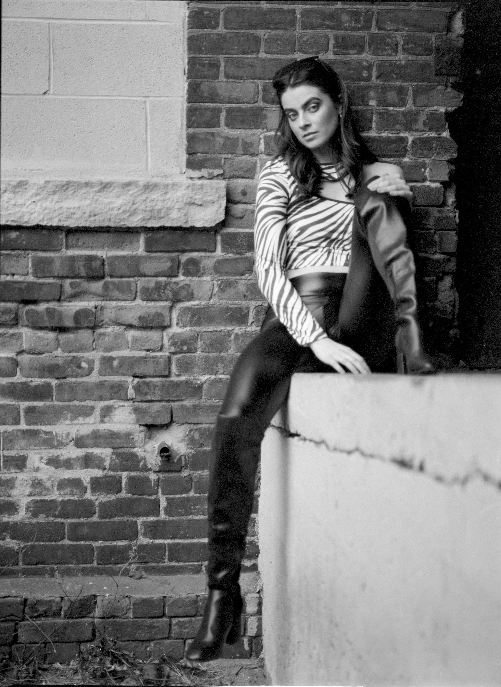 Woman in zebra top and leather pants sits on ledge against brick wall.