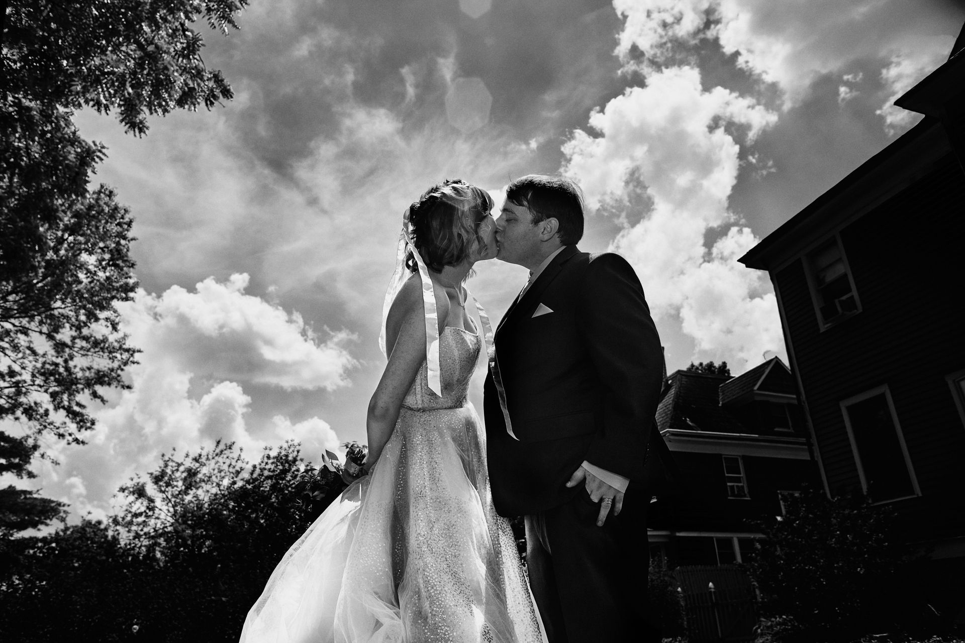 Bride and groom kissing outdoors; sunny sky with puffy clouds; dark house in the background.