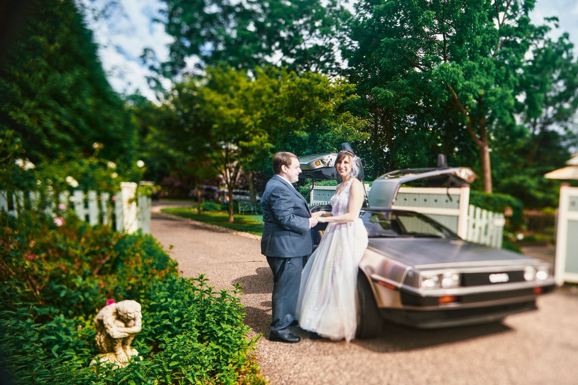 Bride and groom by a DeLorean, the groom in a suit and the bride in a gown. Green foliage and a white fence surround them.