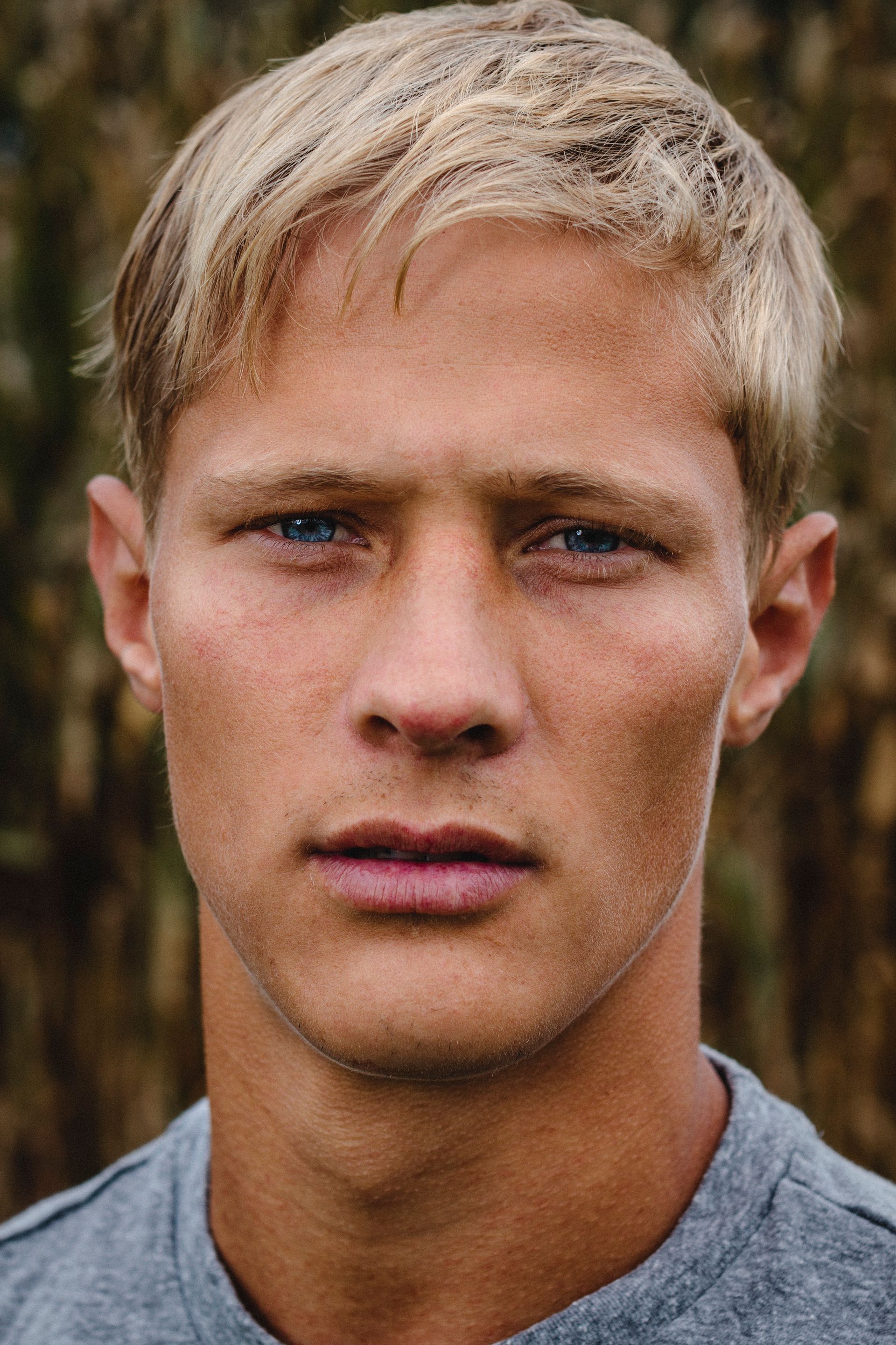 Blond-haired man with blue eyes stares forward. Outdoors, wearing a gray shirt.
