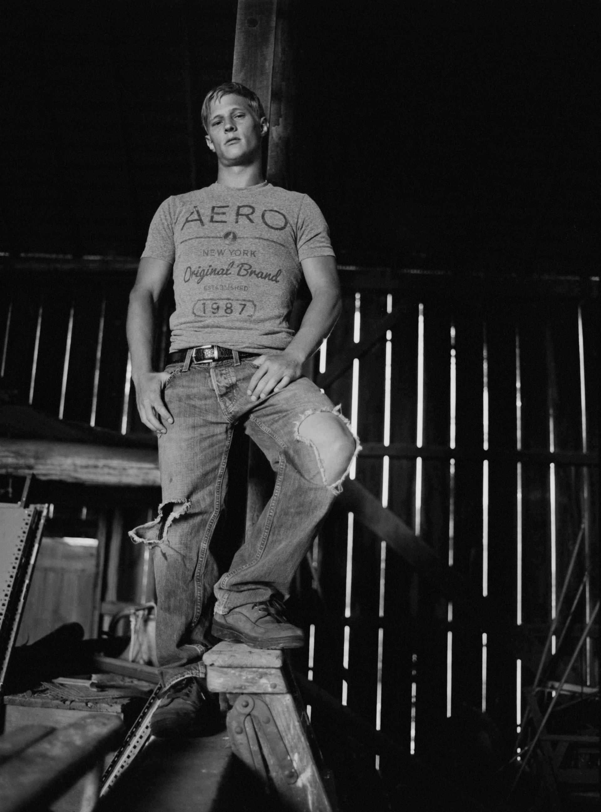 Blonde man in torn jeans and t-shirt poses inside a rustic wooden structure.