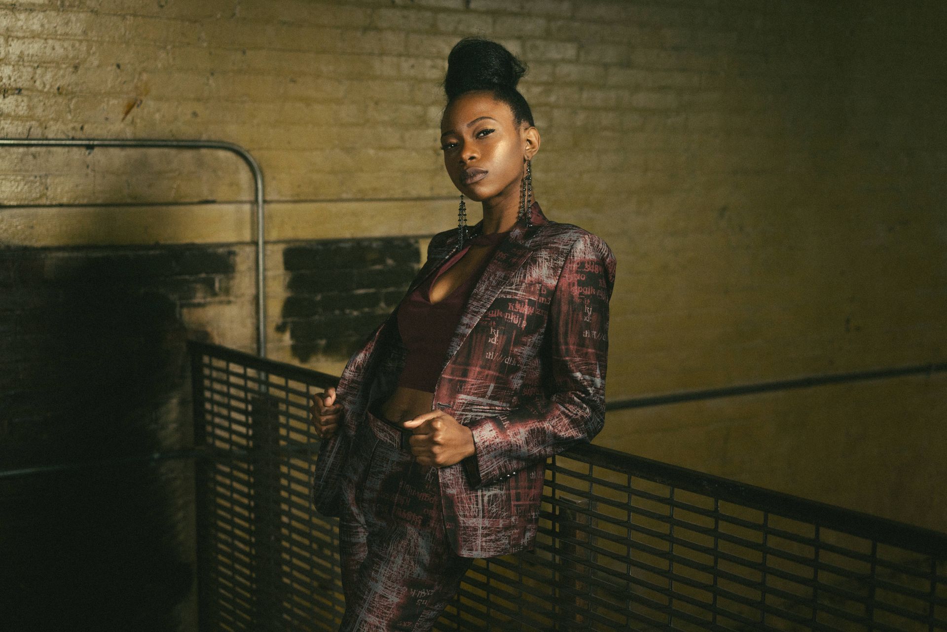 A stylish Black woman leans on a railing, wearing a patterned suit, dark top, and her hair in a bun.
