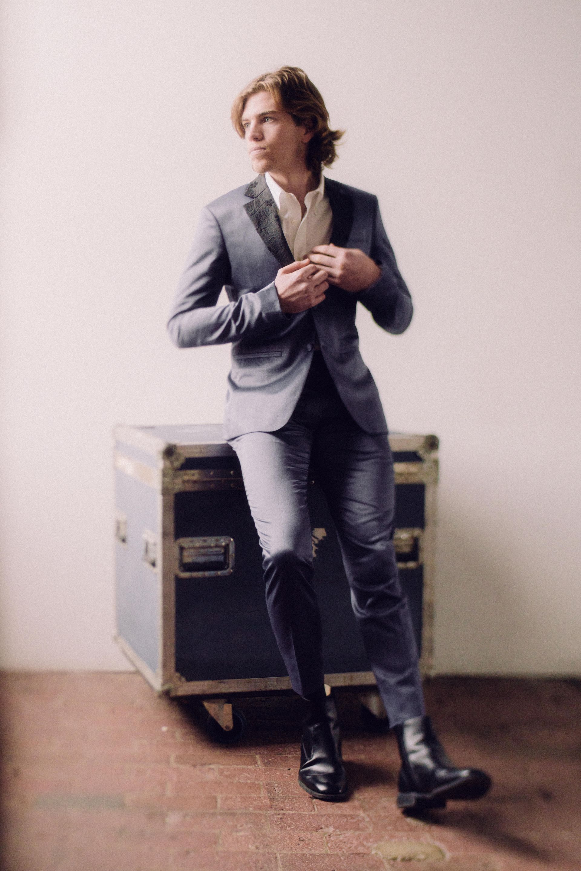 Man in a blue suit leaning on a box, buttoning his jacket. He has long blond hair and is in a studio setting.