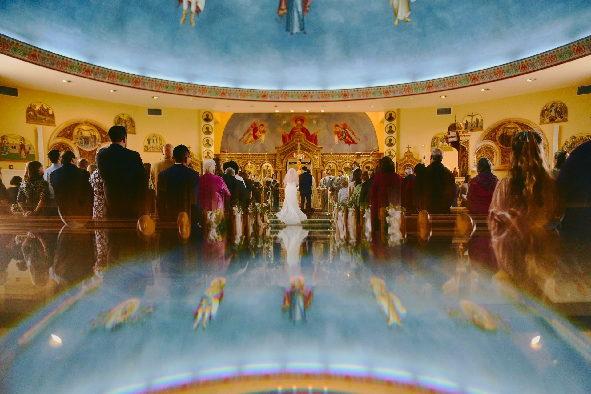 Wedding ceremony in a domed church, bride and groom at altar, guests seated, colorful reflections in foreground.