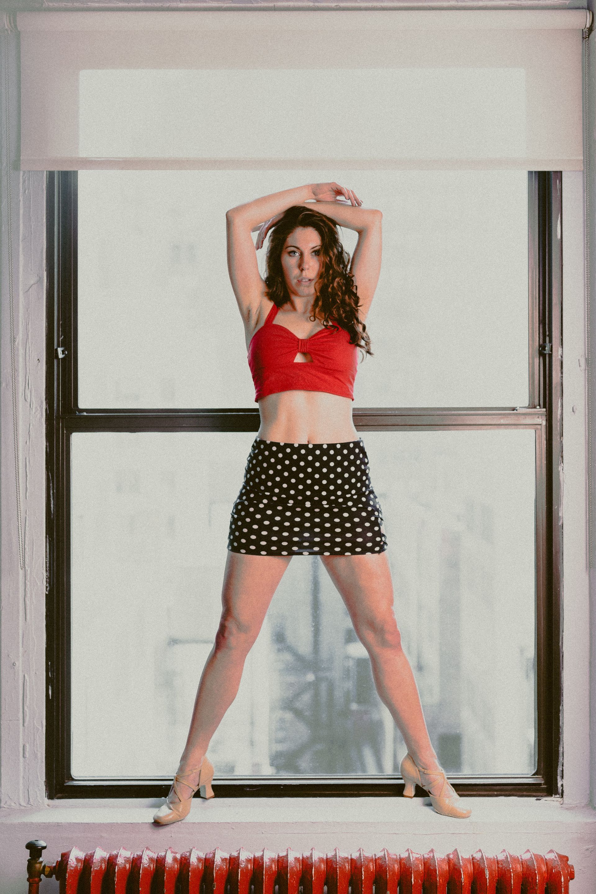 Woman in red top and polka dot skirt, arms raised, posing in front of a window.
