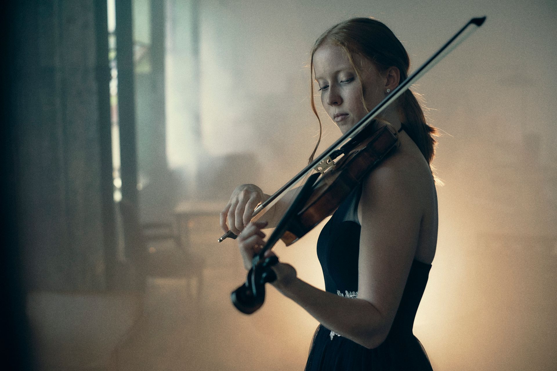 Woman with red hair plays a violin in a dimly lit room, wearing a black dress.