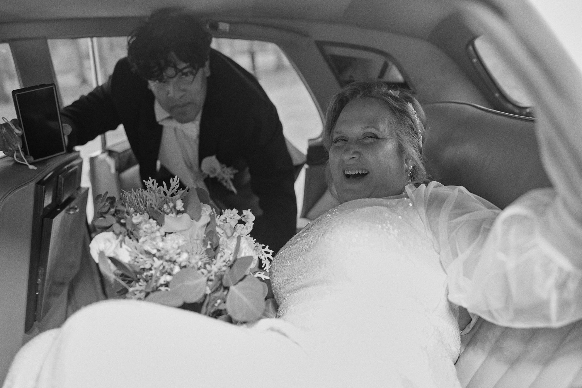 Bride in wedding dress, smiling inside car. Man holding phone, leans in. Bouquet visible.