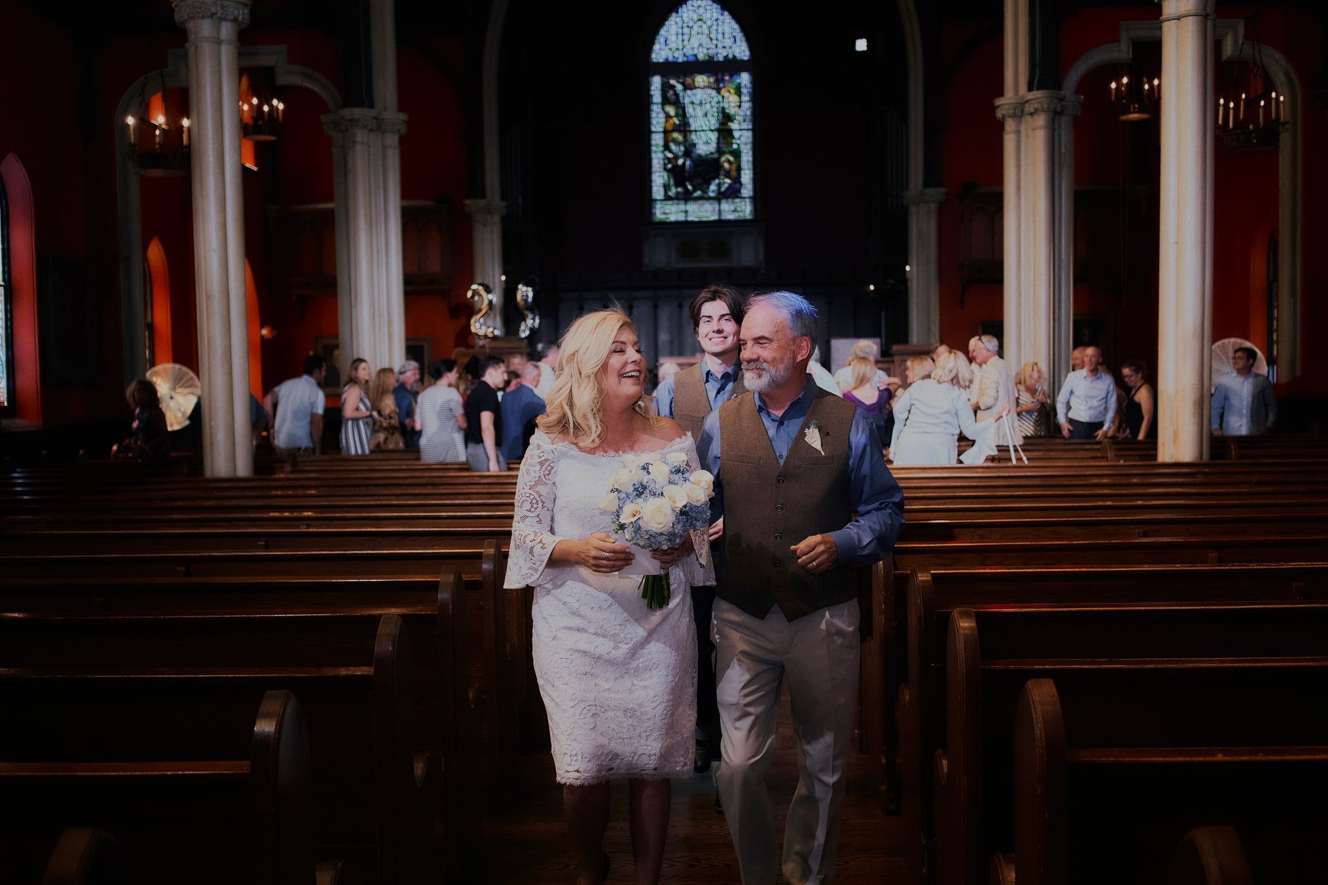 Newlyweds exiting a church, smiling. Bride in white dress, groom in vest. Guests applaud in pews.