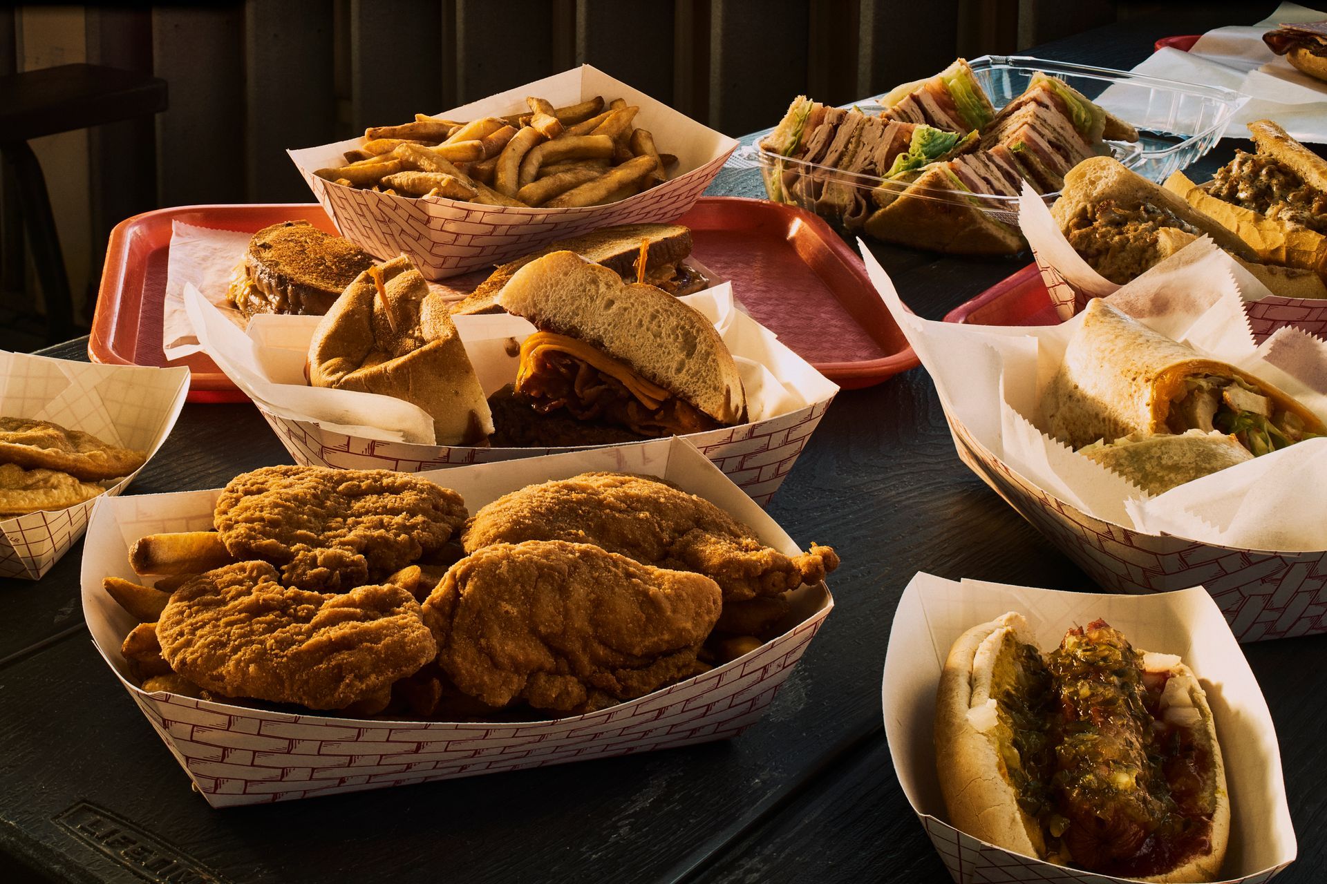 Assorted fast food on a table, including fries, chicken, sandwiches, and a hot dog in paper containers.