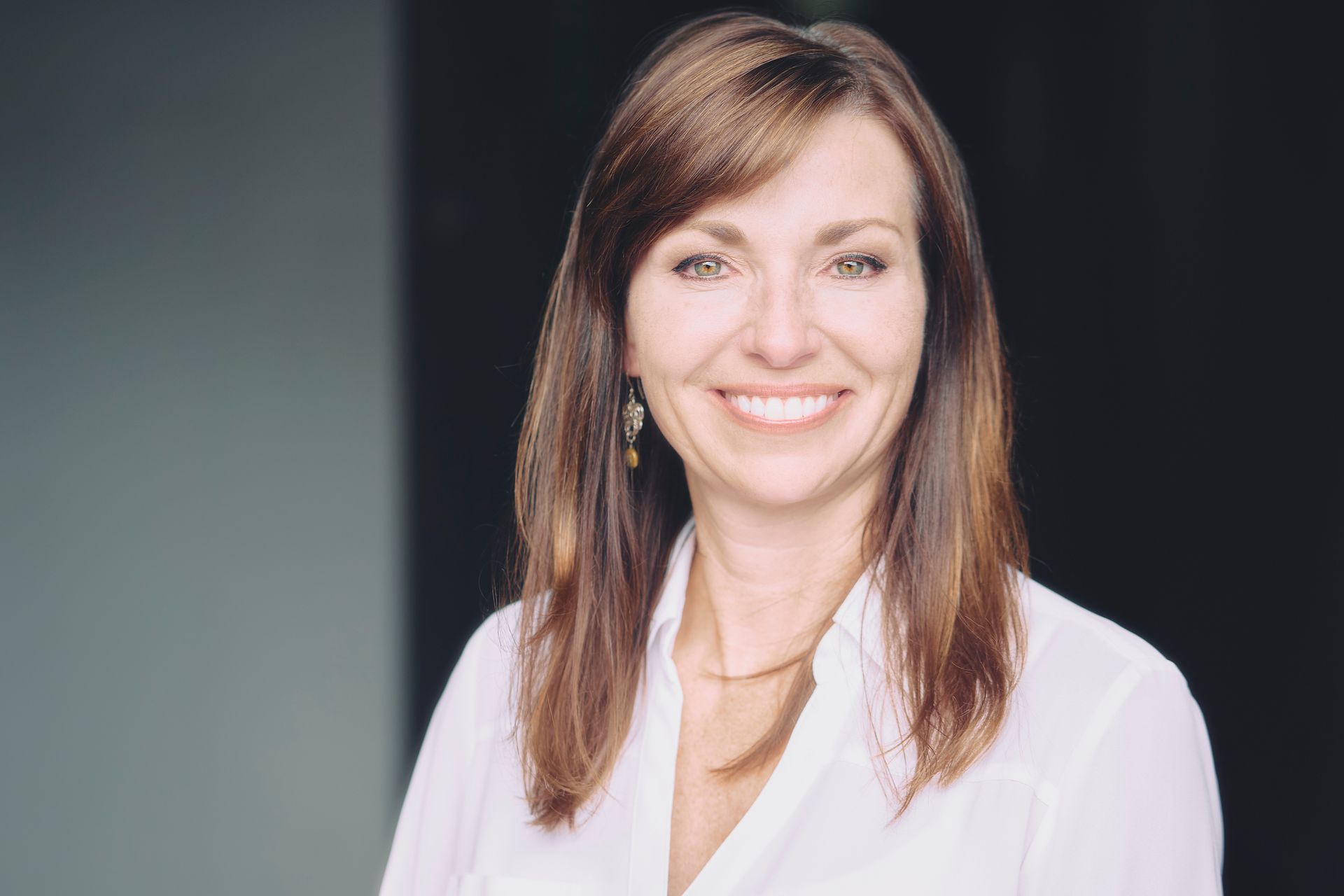 Woman with brown hair and a white shirt smiles against a gray and black background.