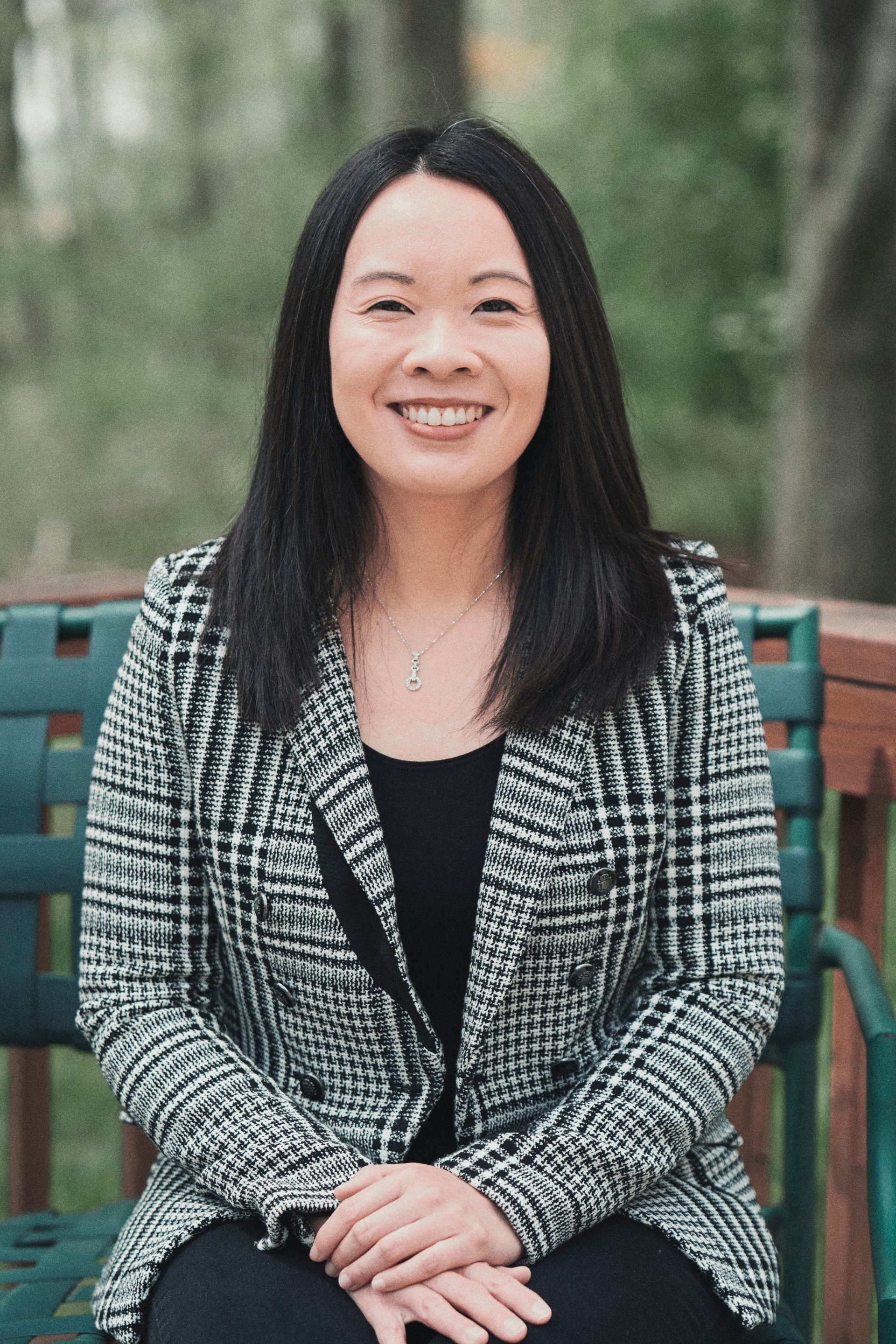 Woman with dark hair, smiling, in a black and white jacket, sitting outdoors.