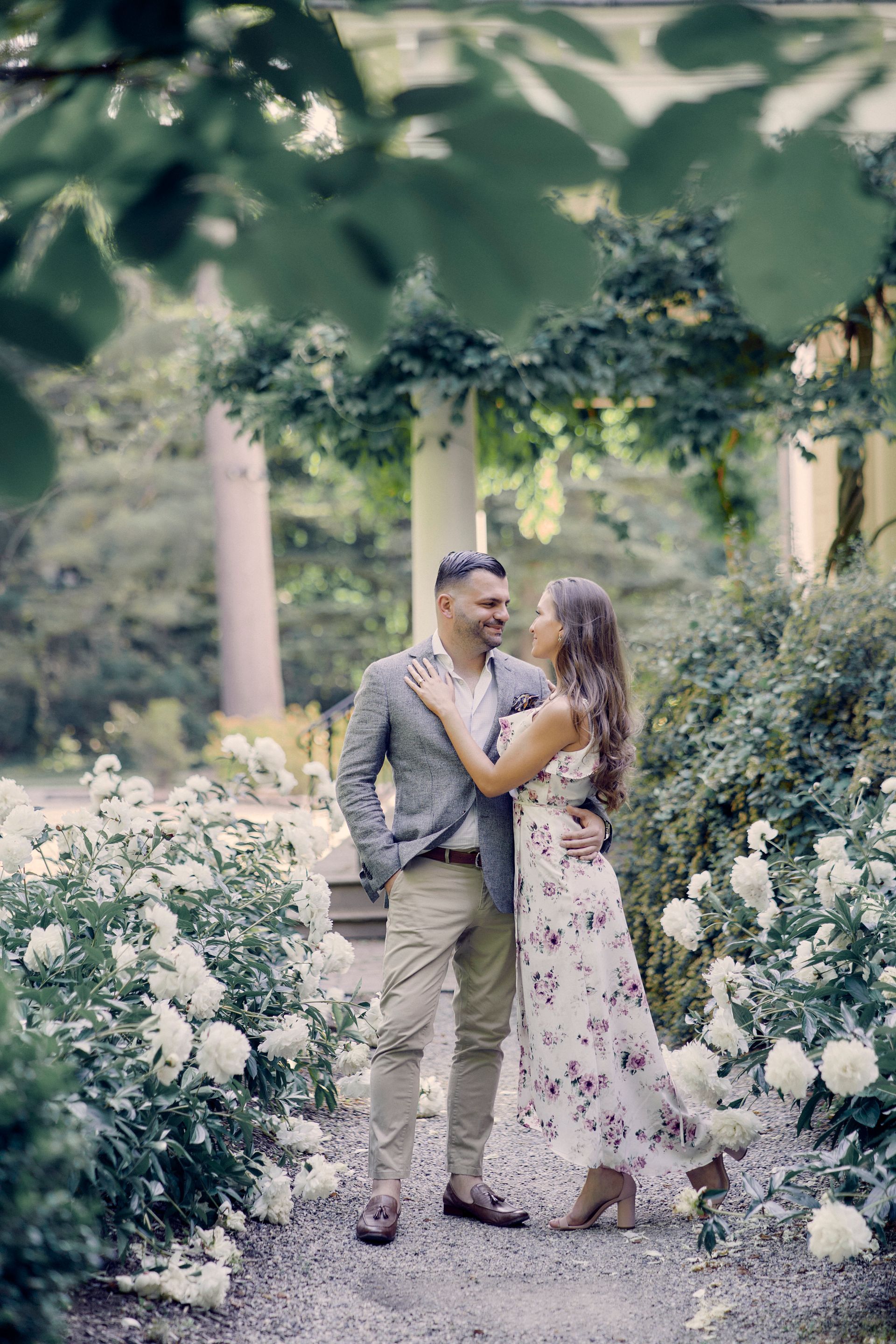 Couple in a garden smiles at each other, surrounded by white flowers and greenery.