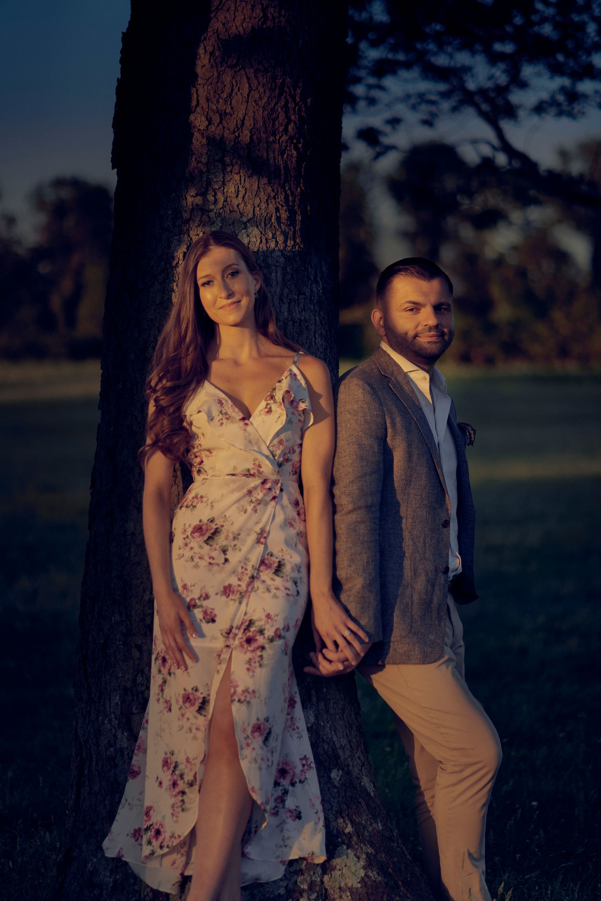 Couple holding hands, standing by a tree. Woman in floral dress, man in blazer, both smiling in an outdoor setting.