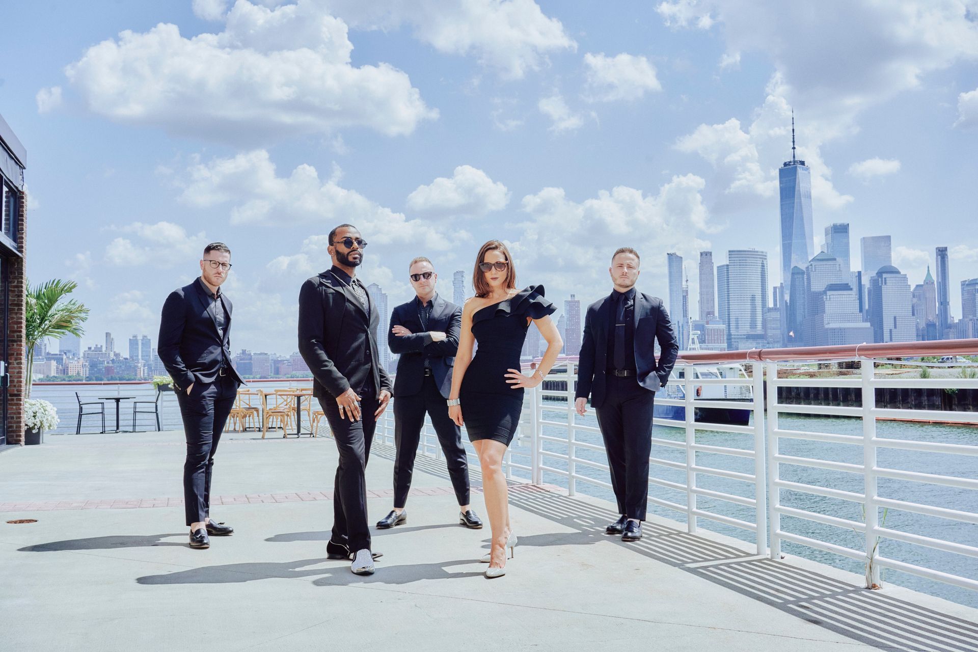 Five people in formal black attire pose by water, NYC skyline in background. Woman in center. Sunny day.