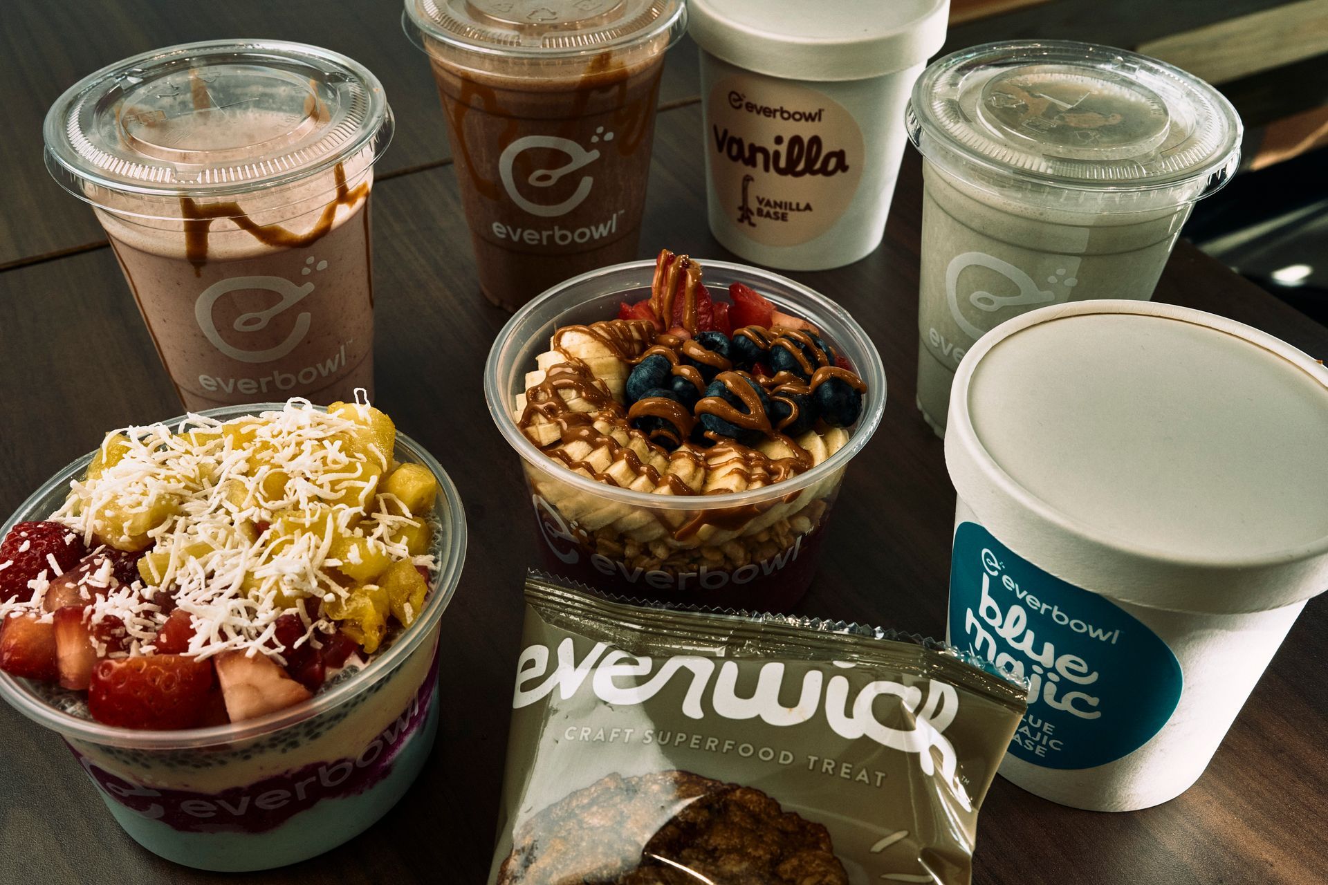 Assortment of Everbowl food and drinks: fruit bowls, smoothies, and a granola bar on a wooden table.