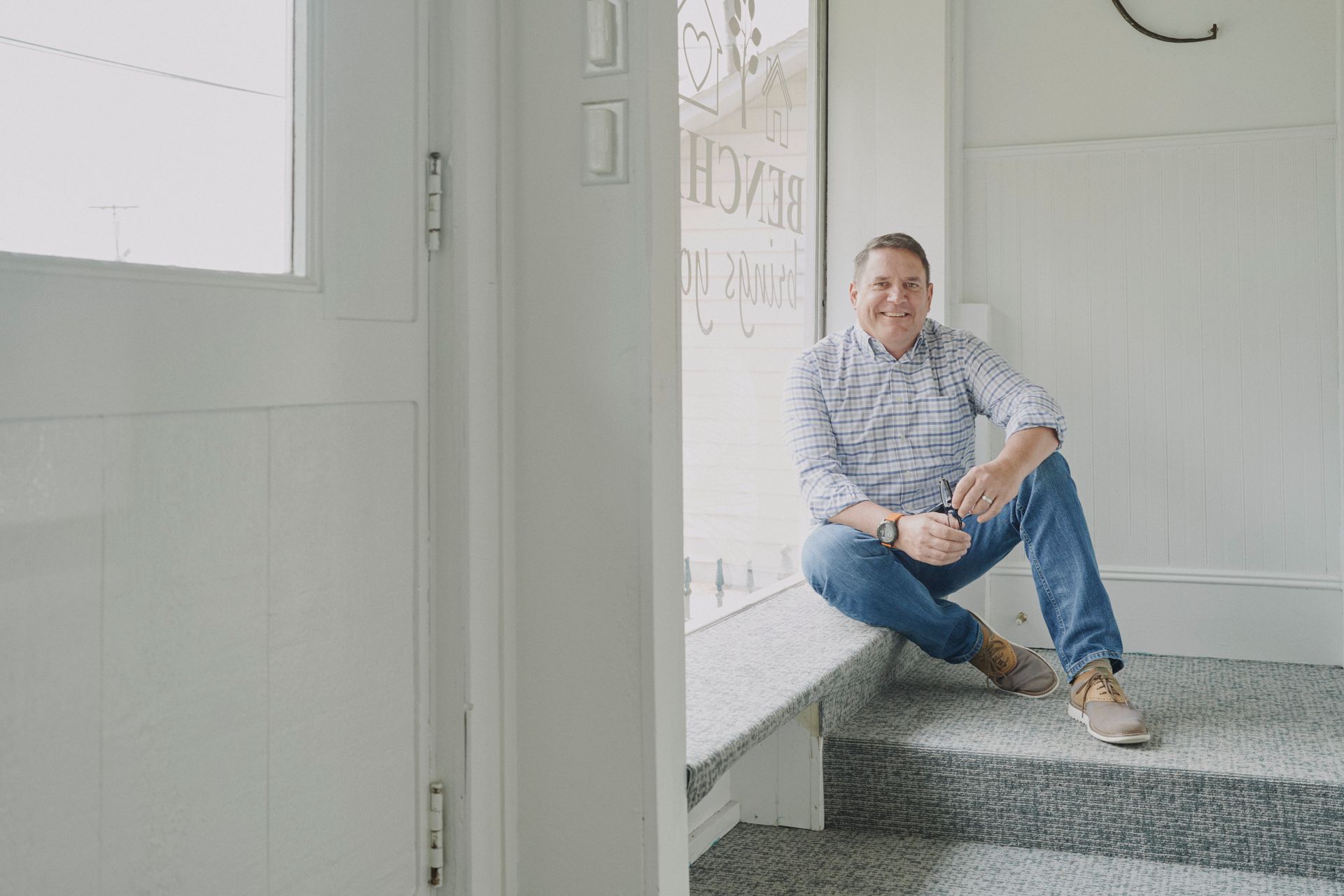 Man sitting on steps, smiling, wearing blue jeans, light button-up shirt and tan shoes.