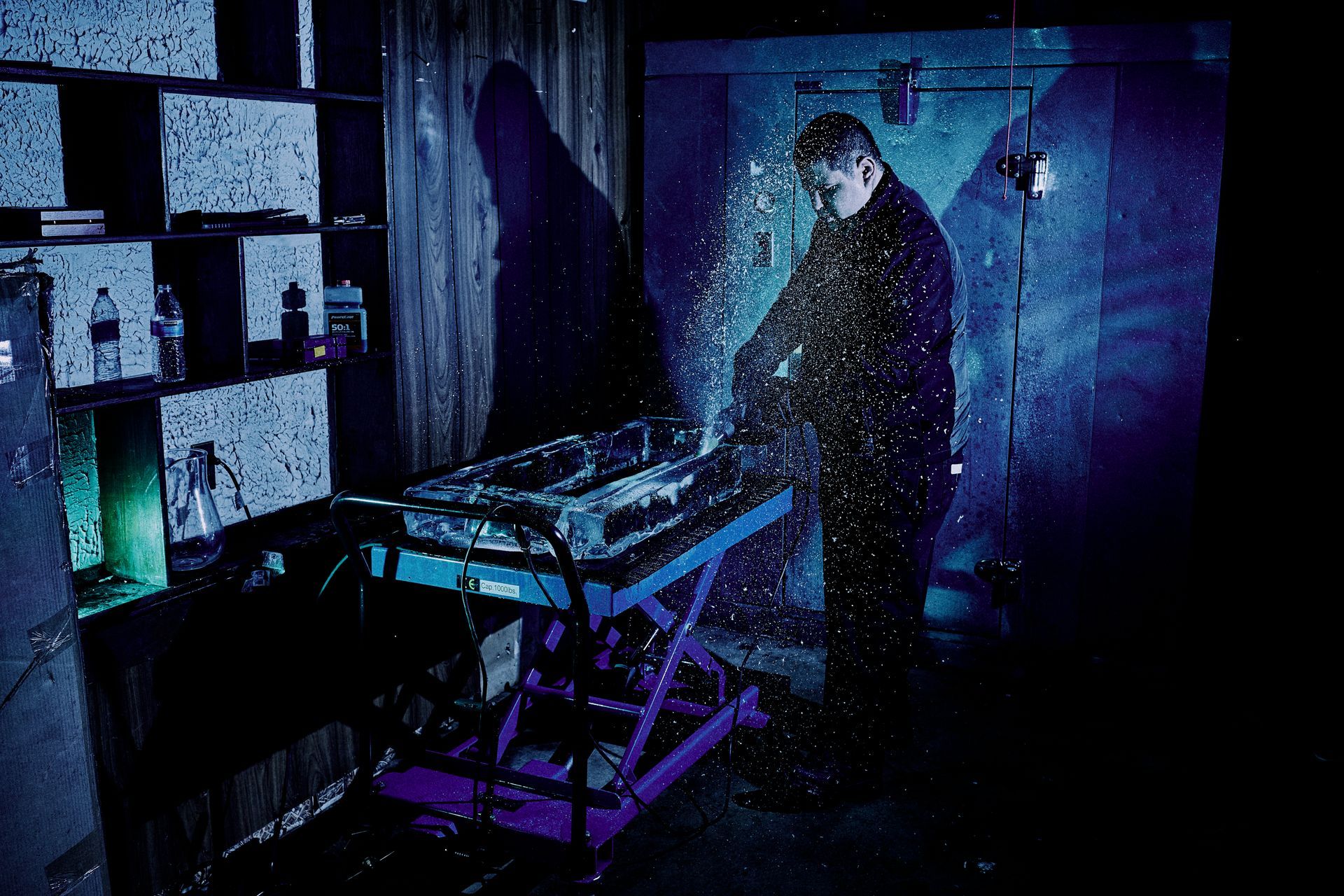 Man standing in a dark, blue-lit room, using a synthesizer. Snow-like effect visible, creating atmosphere.