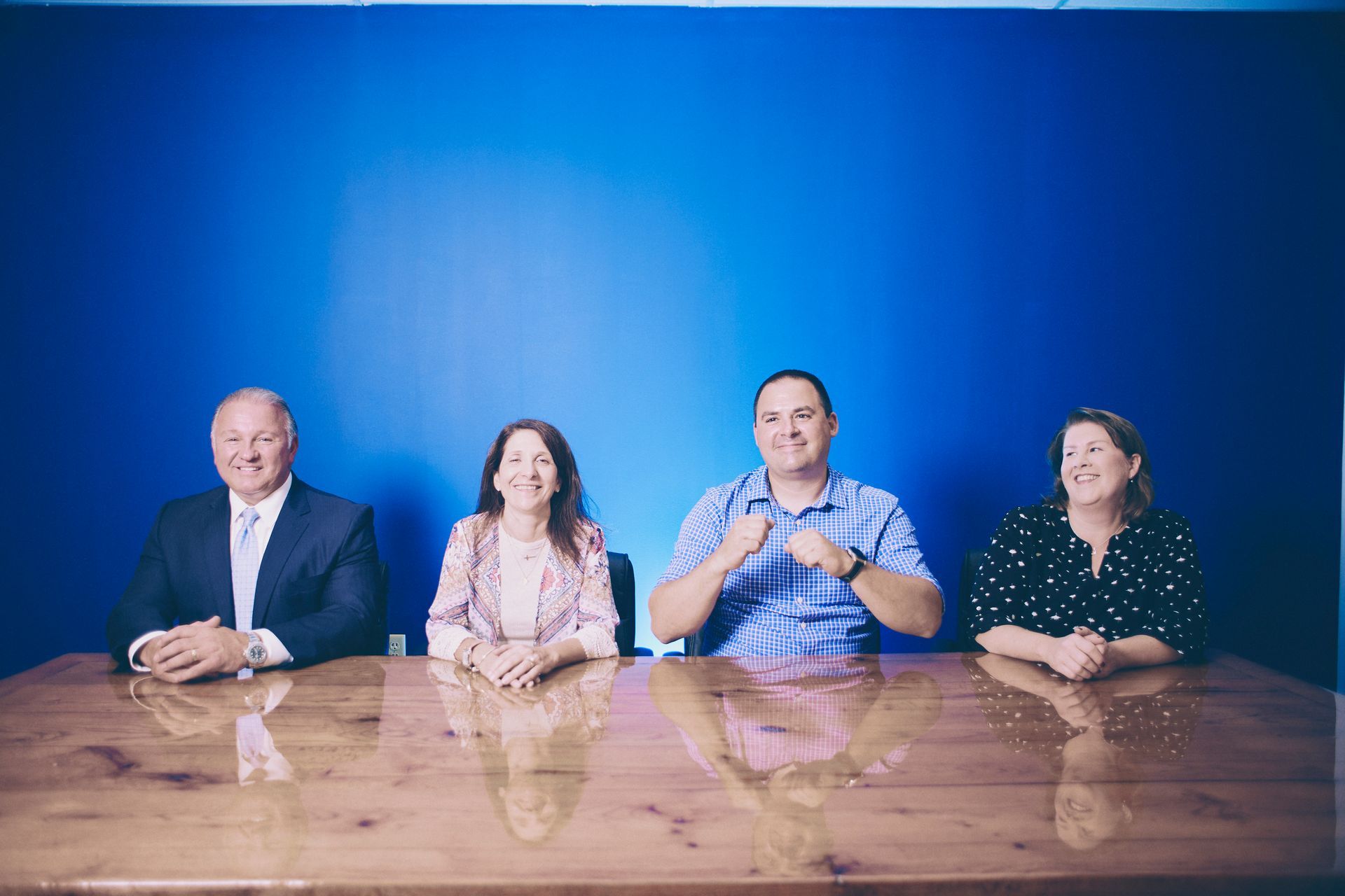 Four people seated at a wood table against a blue wall. Man in the middle is making a fist.