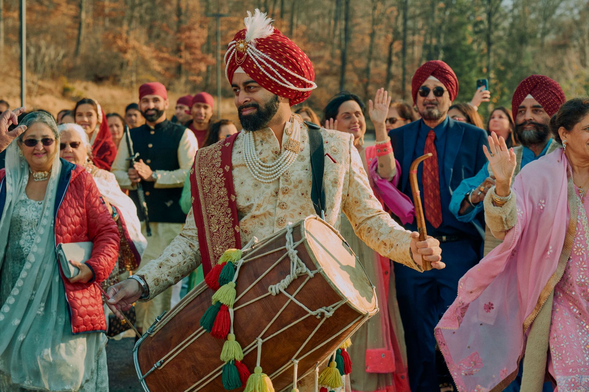 Punjabi man playing a dhol drum, leading a wedding procession outdoors; many people waving.