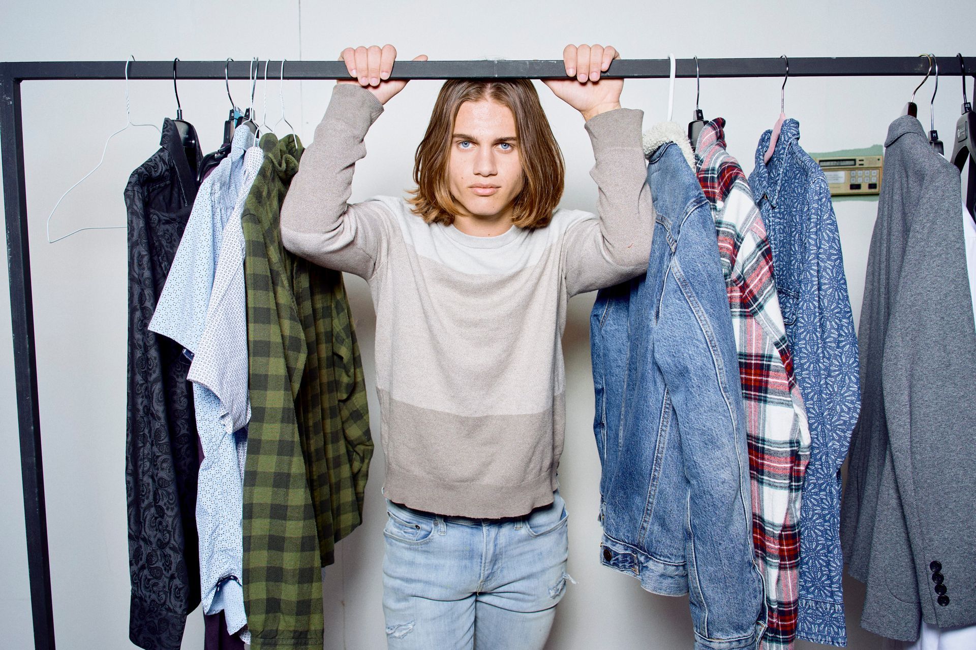 Young person with long hair, wearing a sweater, holding onto a clothing rack, surrounded by hanging clothes.