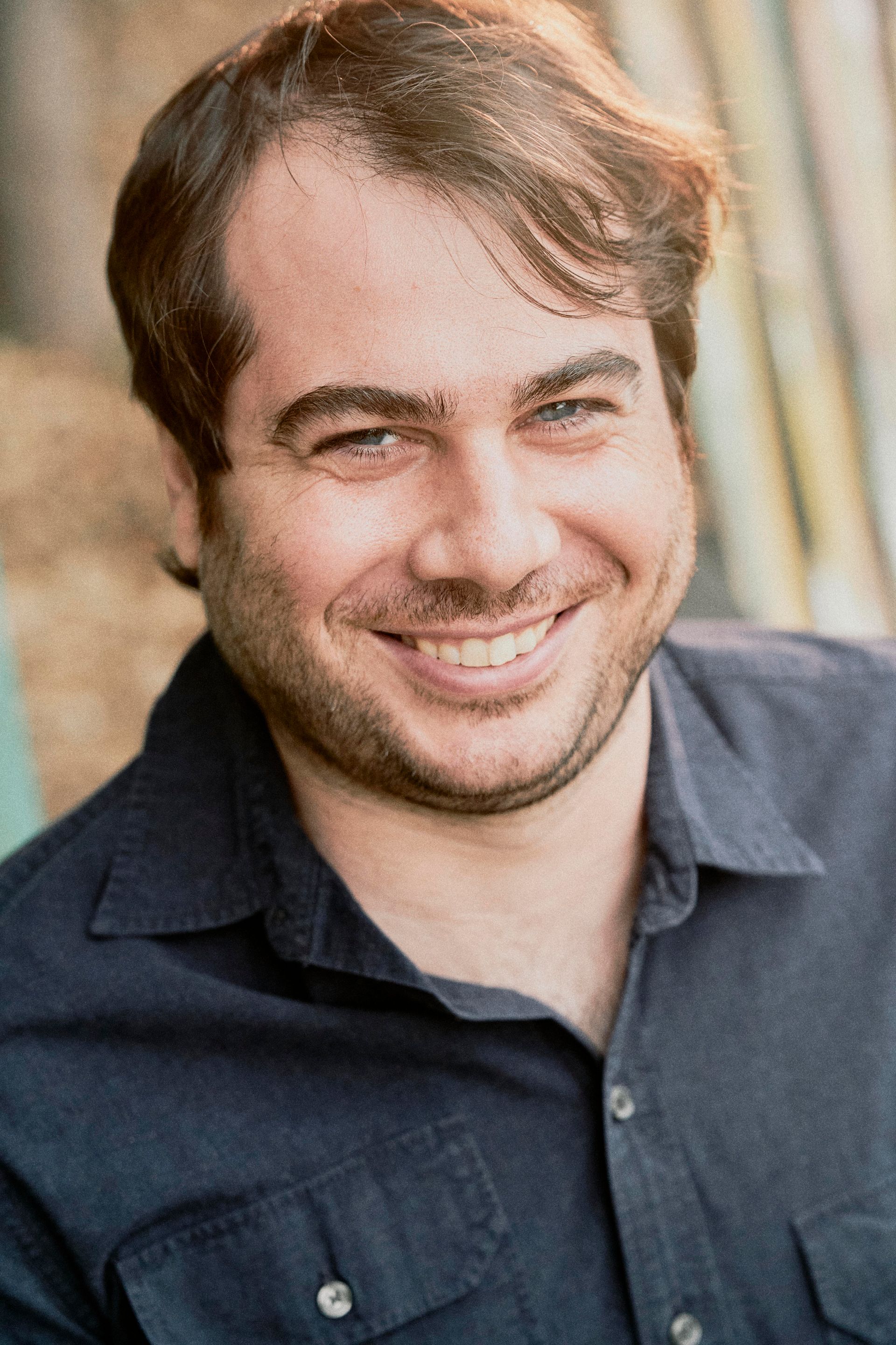 Man with brown hair, smiling, wearing a dark blue button-down shirt, set outside.
