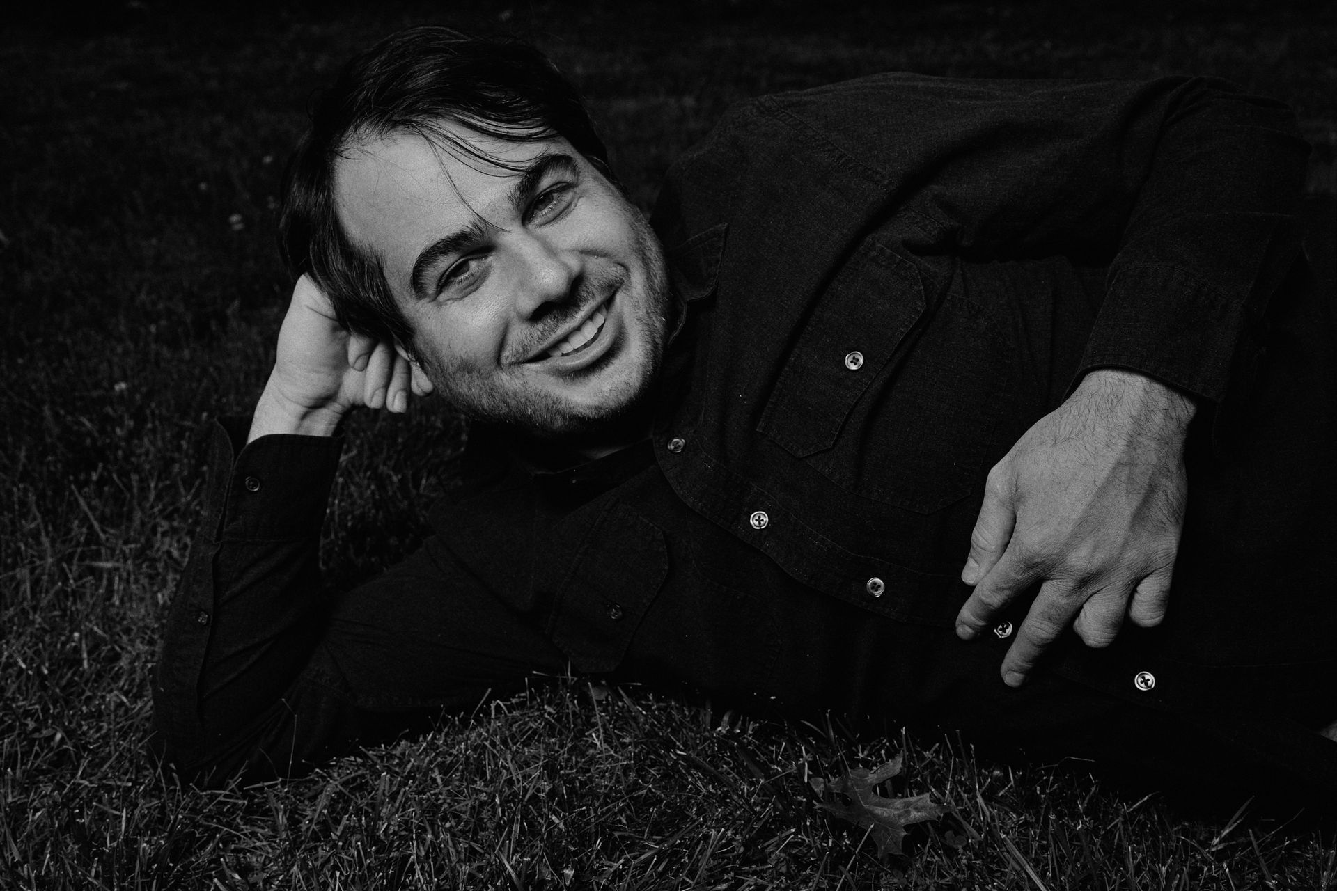 Man lying on grass, smiling, black shirt, arm under head, black and white photo.