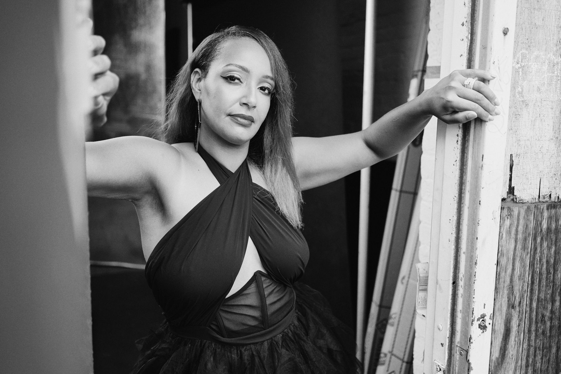 Woman in a black halter dress poses in a doorway, leaning against the frame. Black and white photo.