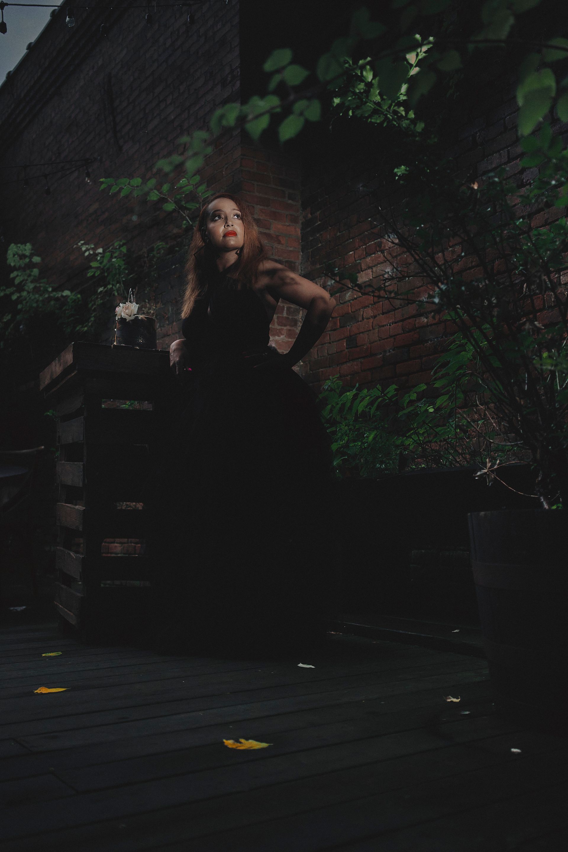 Woman in a black dress posing on a dark patio; brick wall and foliage behind her.