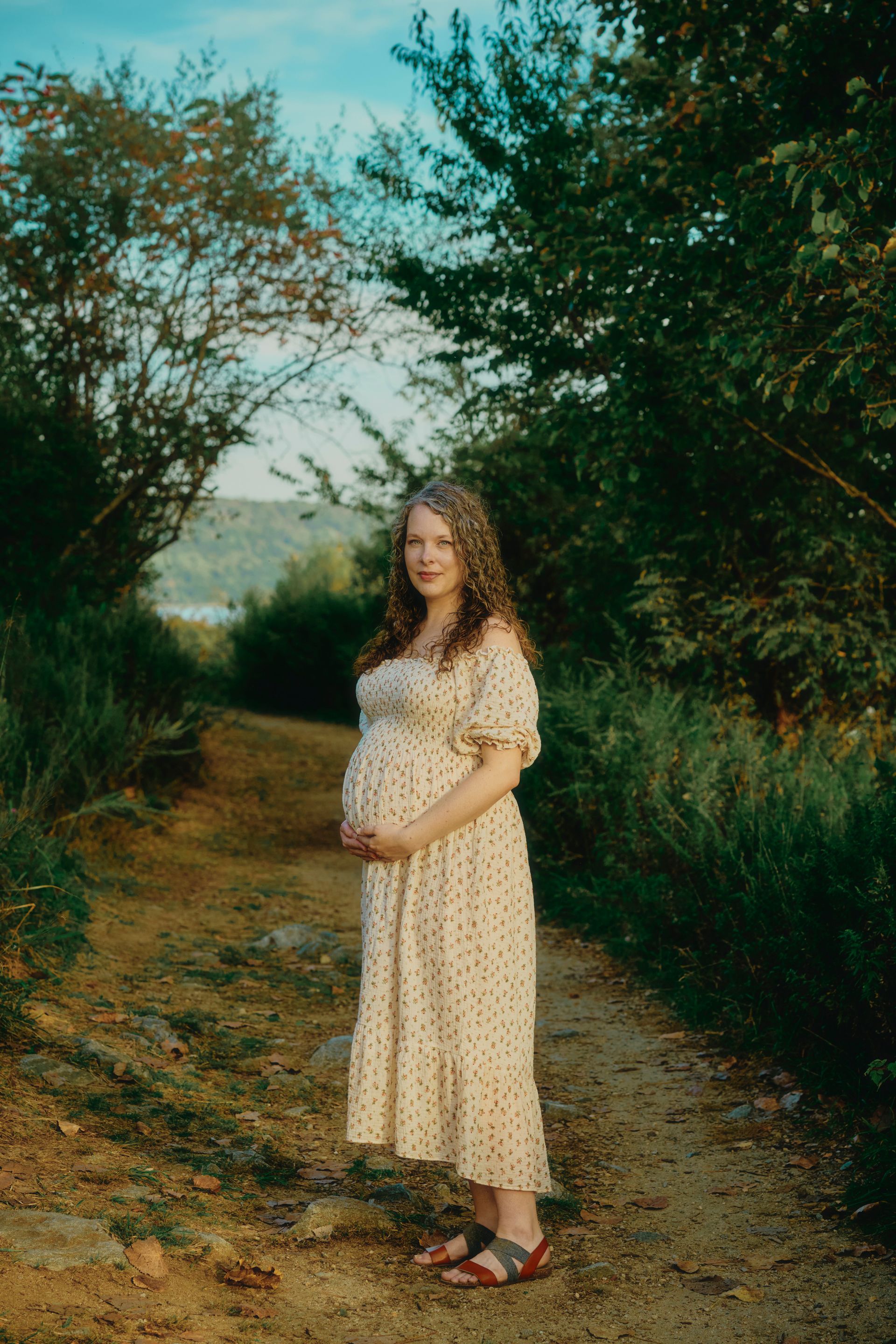 Pregnant person stands in water, sunlight shining. Wearing a white dress, hands on belly.