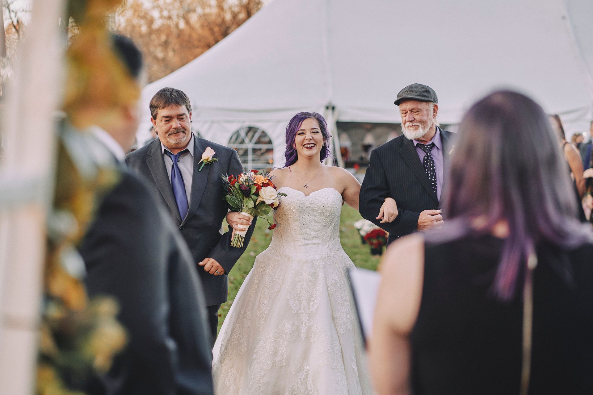 Bride walking down the aisle with two men, smiling, in front of a white tent.