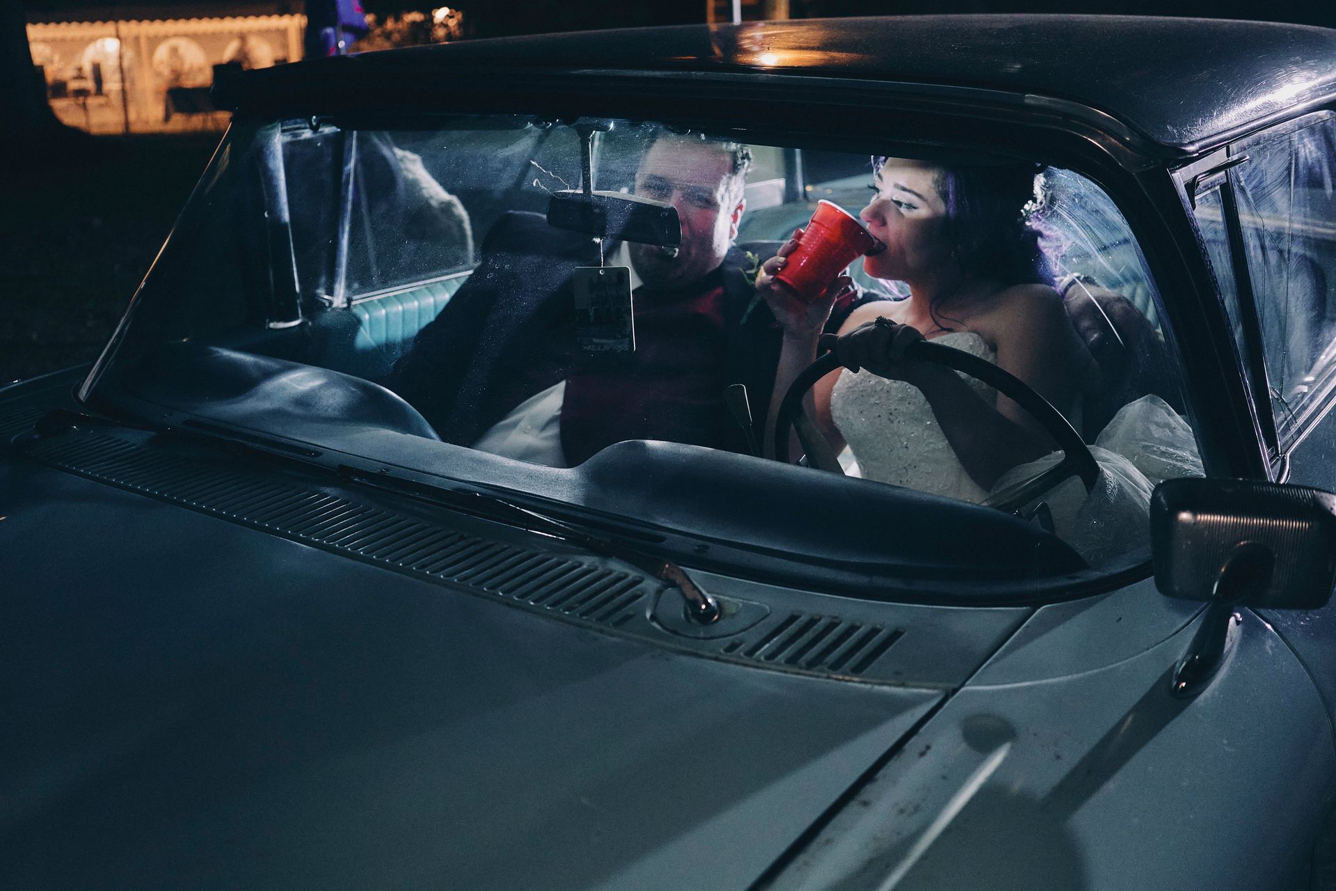 Couple in a vintage car at night, woman drinking from a red cup, man looking at something.