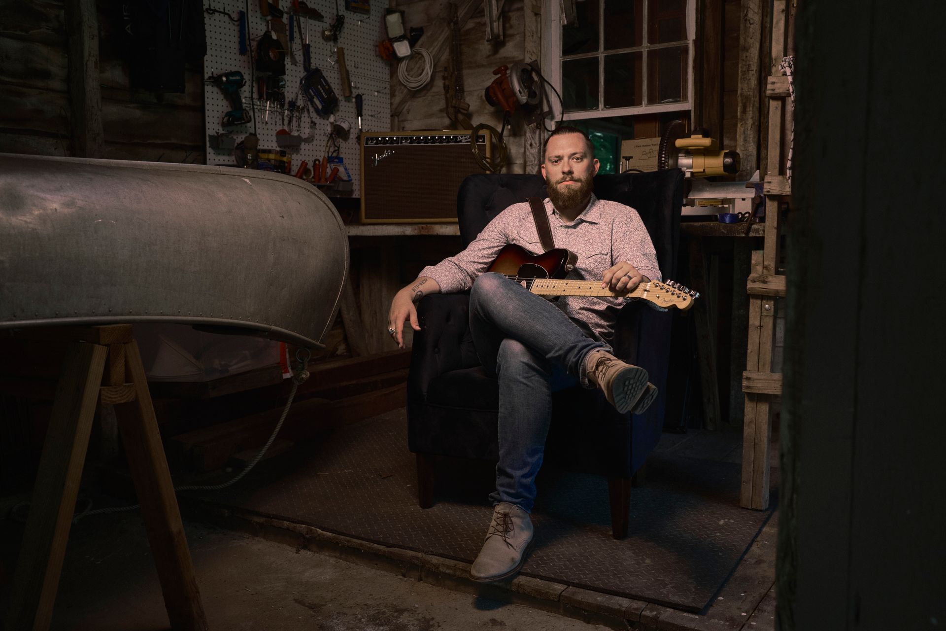 Man in a garage, sitting in a chair with a guitar. Crosses legs. Dark setting.