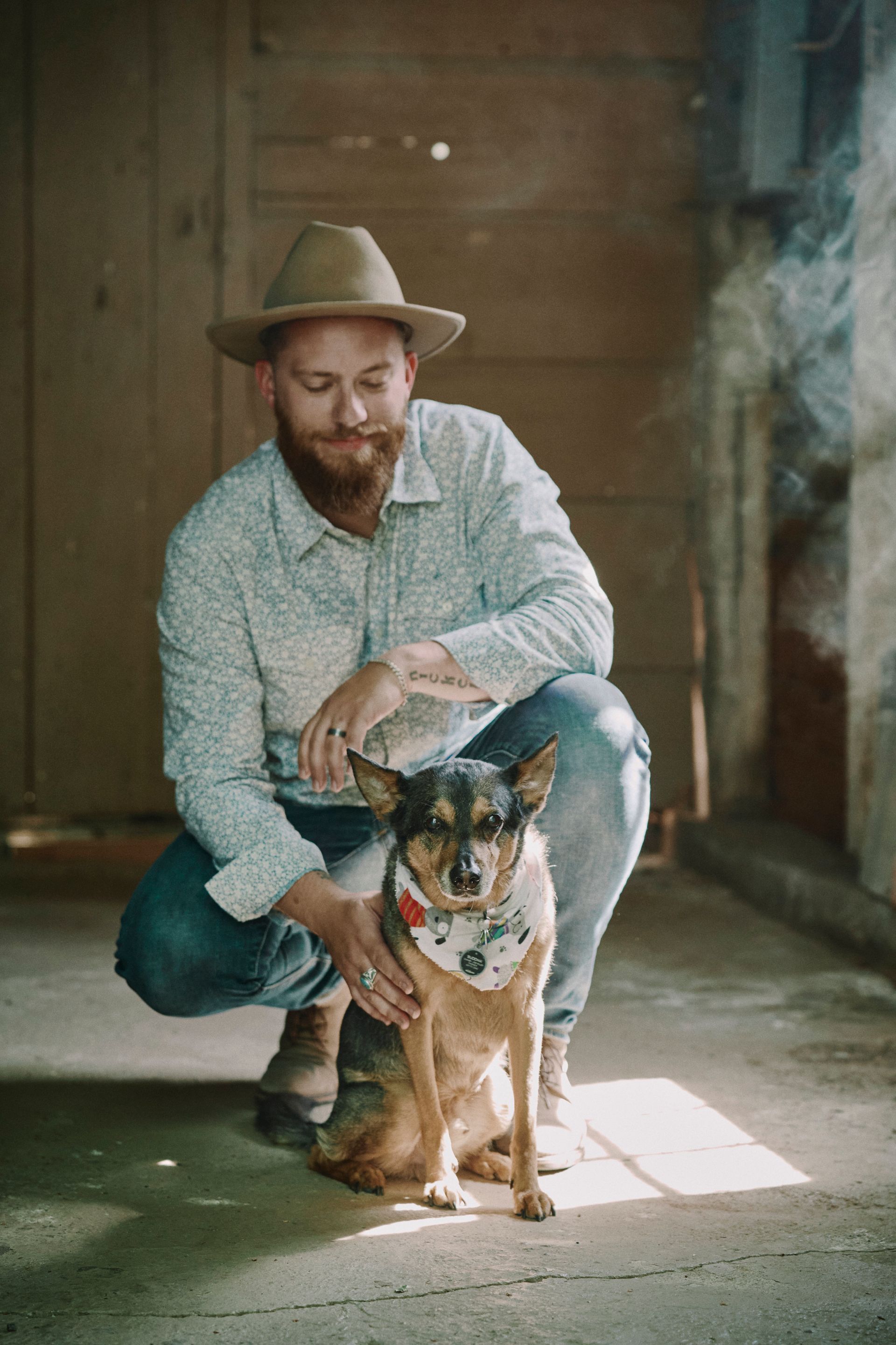 Man in hat squats, petting dog with bandana in rustic doorway.