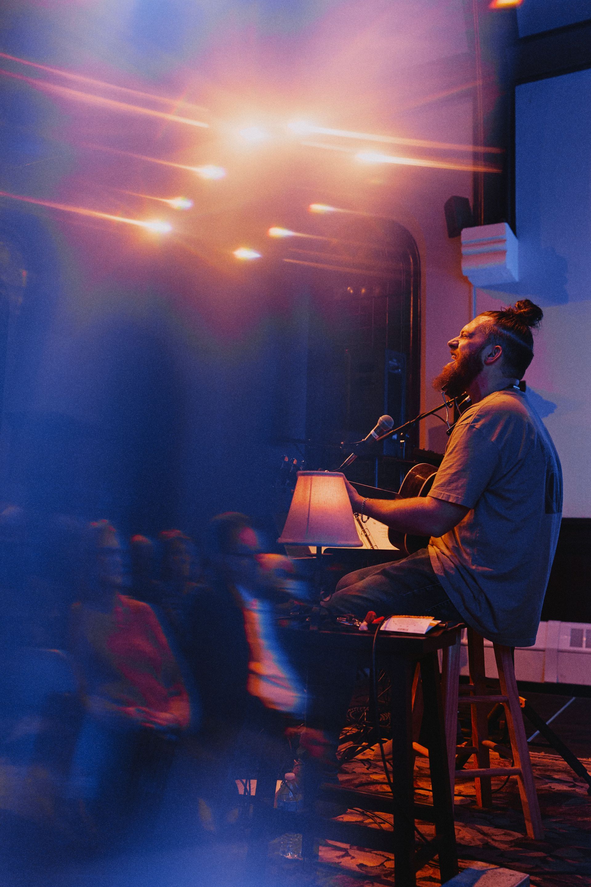 Man with a bun sings at a piano in a dim venue, lit by colorful stage lights.