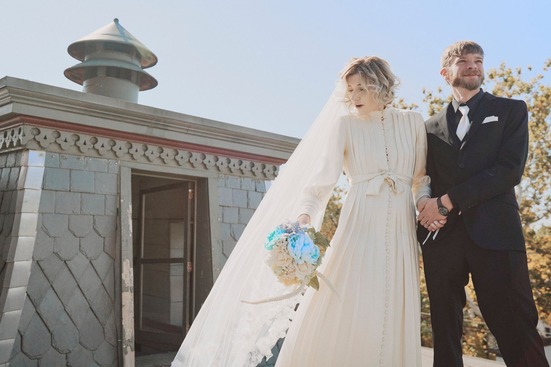 Bride and groom on a rooftop, holding hands. Bride in long white dress, veil, and bouquet. Groom in suit. Sunny.